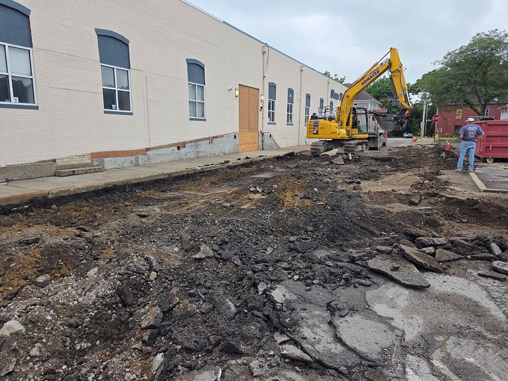 Construction site: Excavator tearing up asphalt next to a light-colored building. Worker stands nearby.