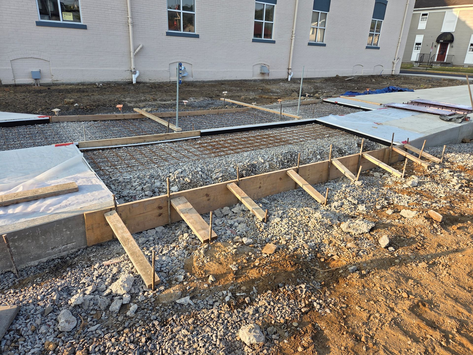 Construction site with wooden forms, gravel, and concrete, in front of a building with windows.