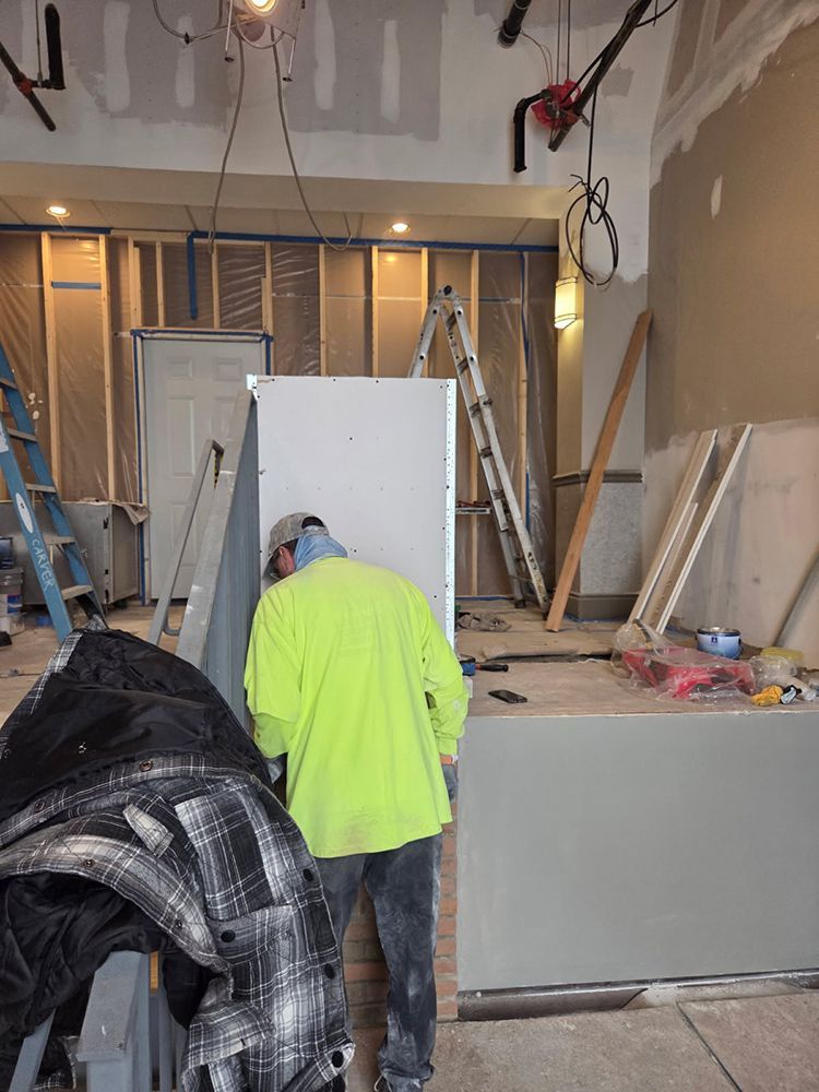 Construction worker wearing a yellow safety shirt working on a drywall installation in a building.