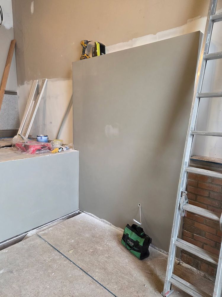 Interior construction scene. Gray drywall against a wall. Tools, a ladder, and materials are visible.