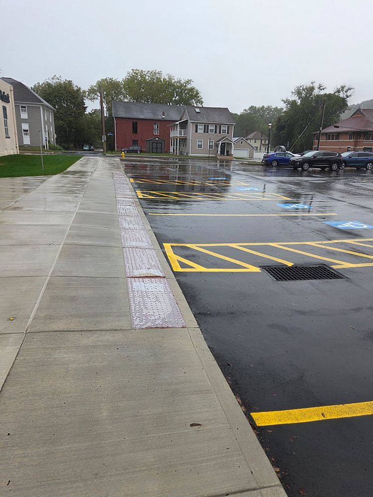 Wet parking lot and sidewalk on a rainy day. Buildings, yellow parking lines, and a handicap space are visible.