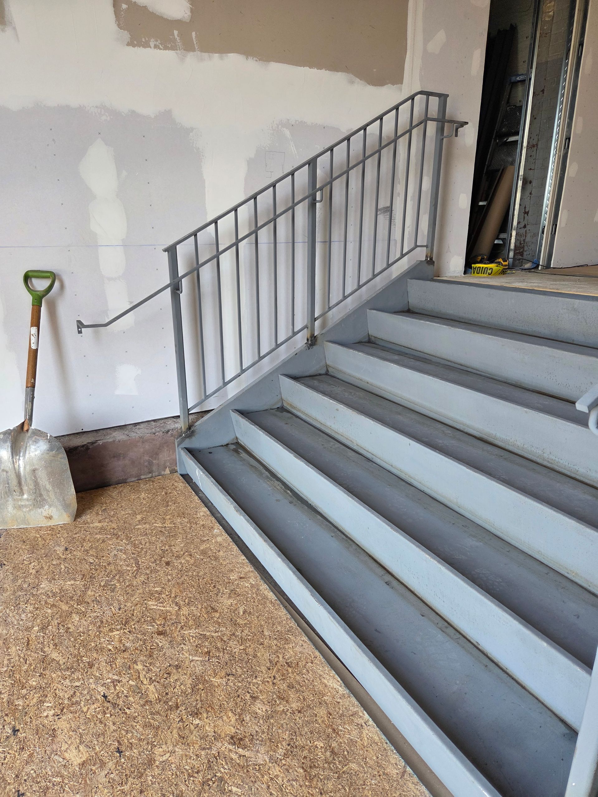 Gray stairwell with metal railing and a shovel leaning against a wall during construction.