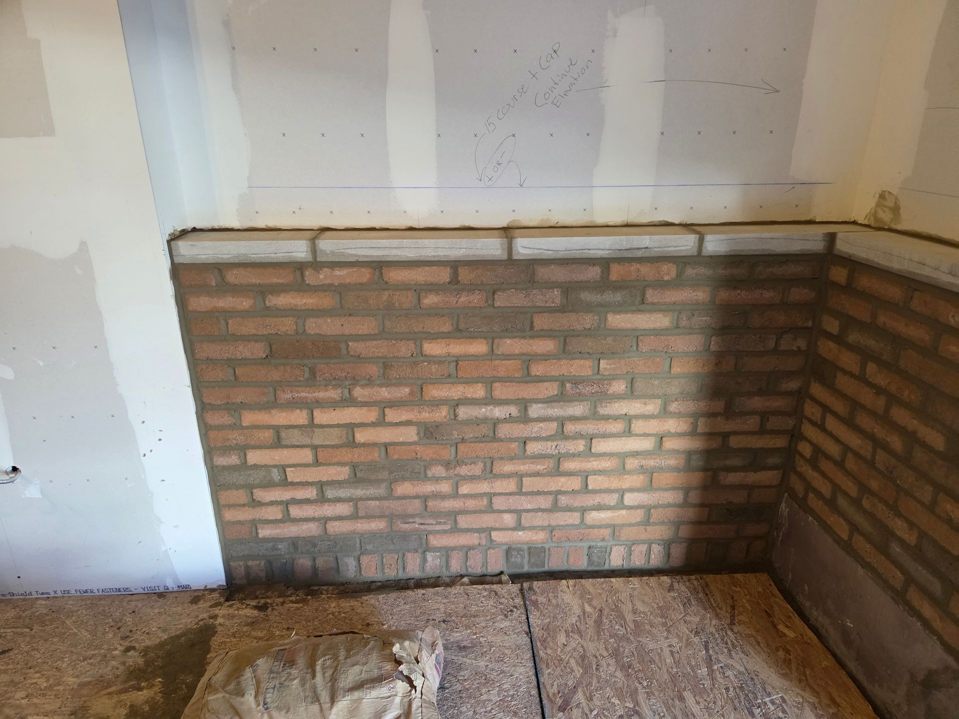 Brick wall construction in progress; interior corner with drywall, red-brown bricks, and mortar.