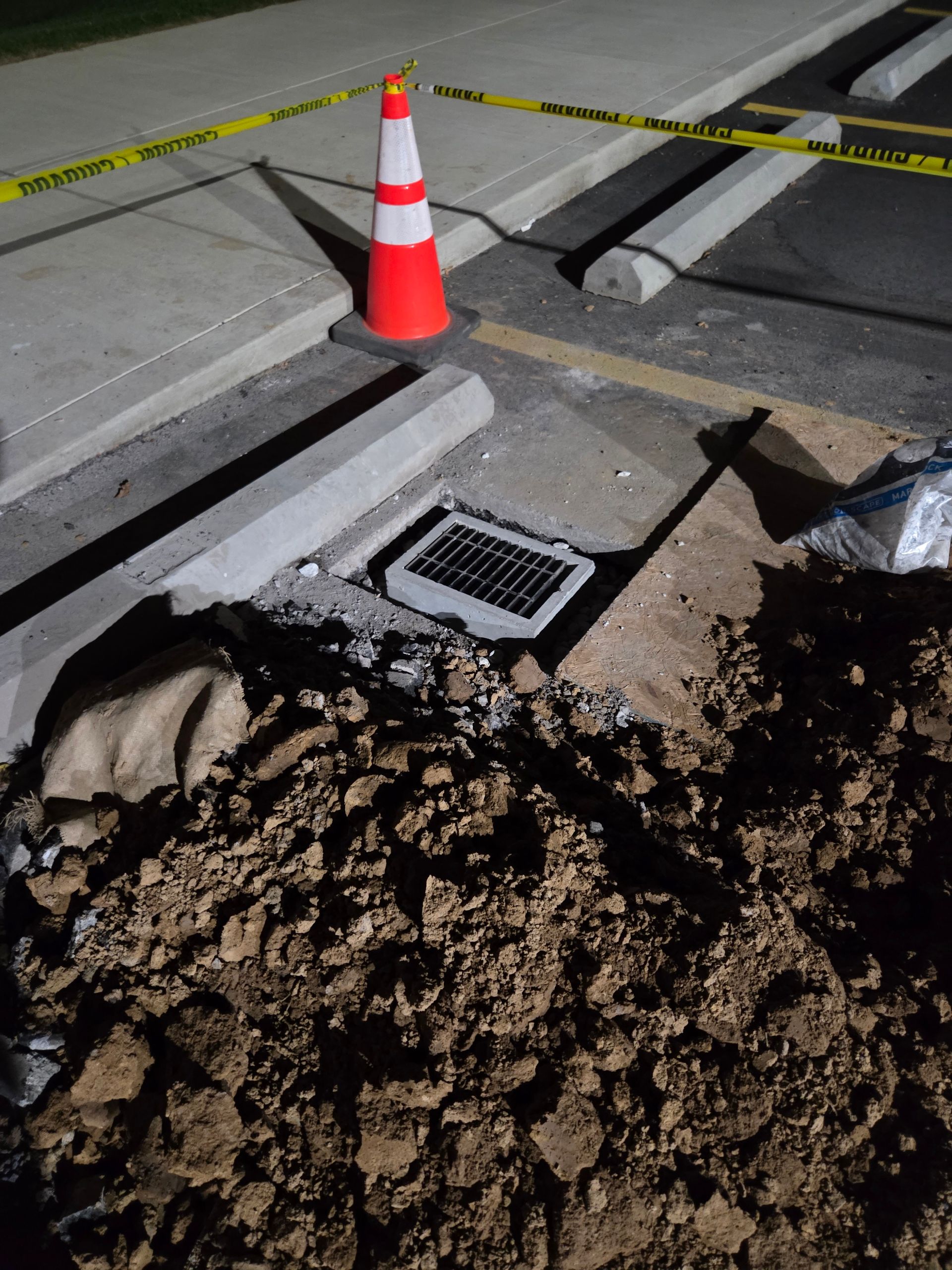 Construction site: broken concrete, dirt, solar panel, orange cone, caution tape in parking area.