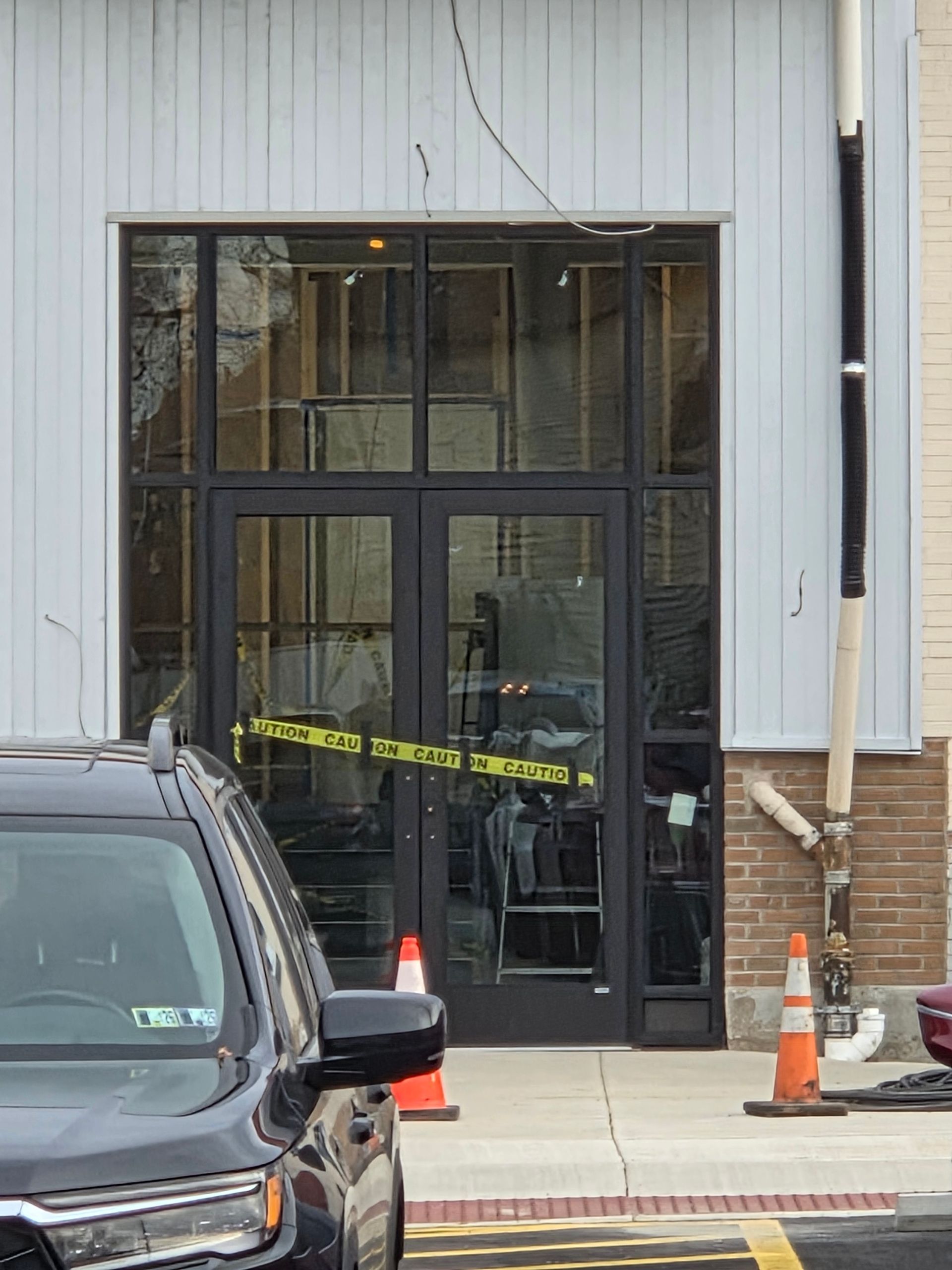 Building entrance with broken glass, caution tape, and a black SUV in front. Orange traffic cones present.