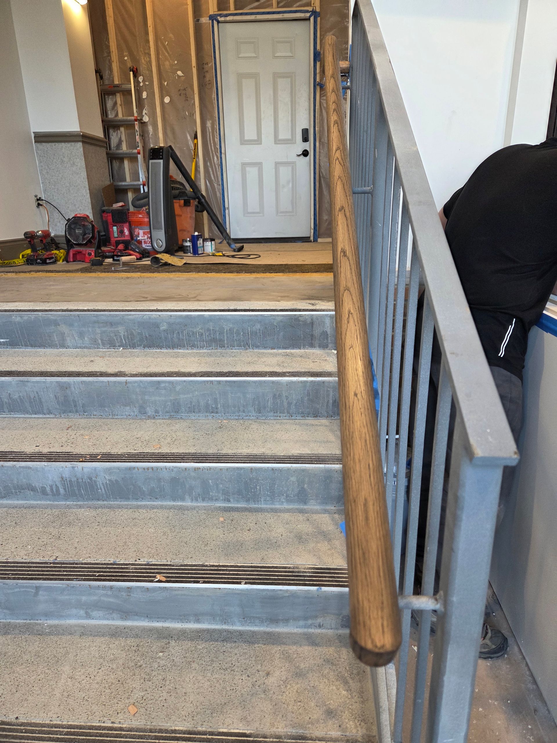 Staircase with metal railing and wooden handrail. Person in black clothing stands nearby. Door in background.