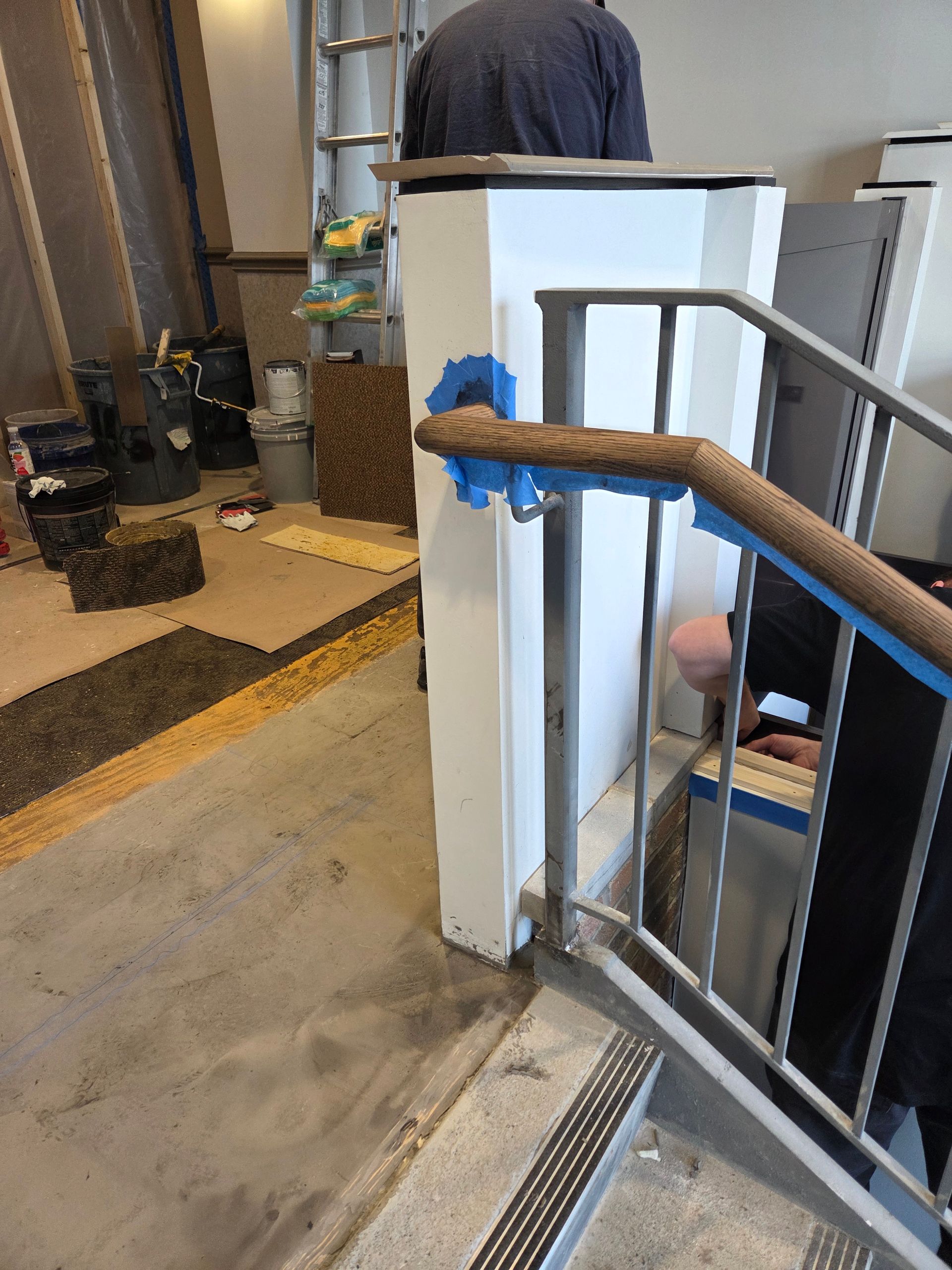 Staircase with wooden handrail, leading up. Handrail meets white pillar, blue tape visible. Person in background.