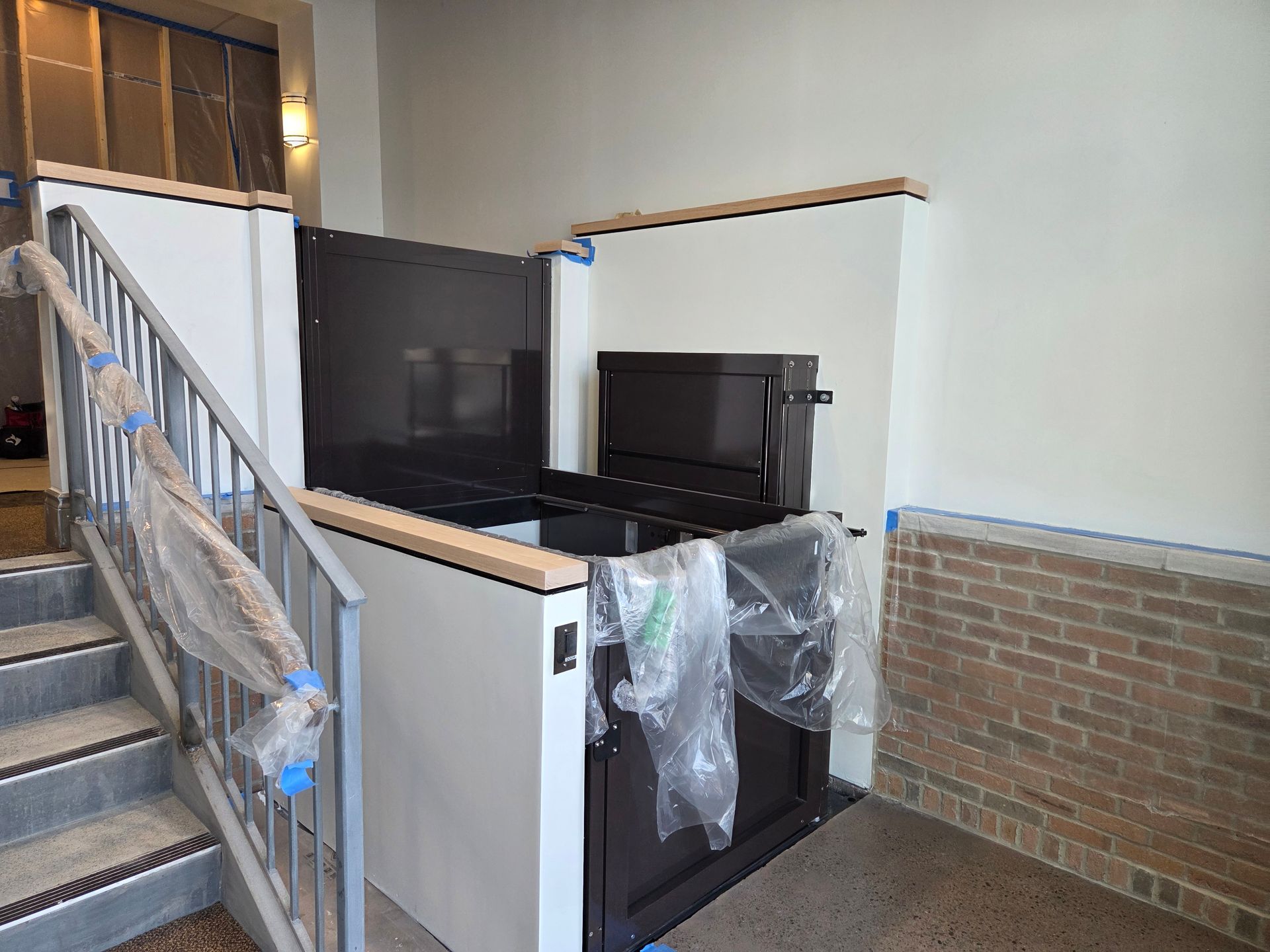 Wheelchair lift installed next to a staircase. The lift has a black door and is encased in white panels with wooden trim.