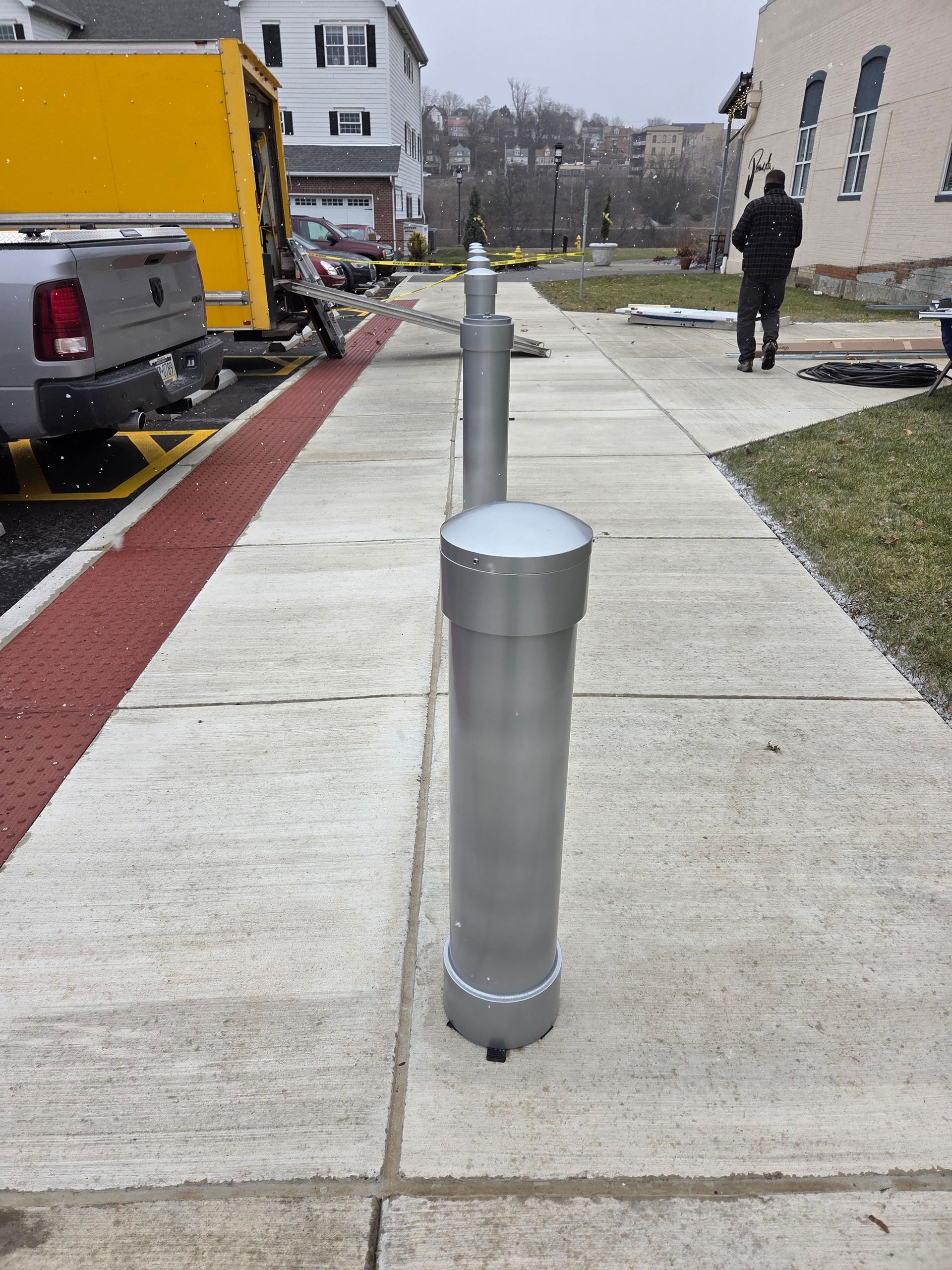 Silver bollard on a sidewalk, adjacent to a parked truck and building; a person walks in the distance.