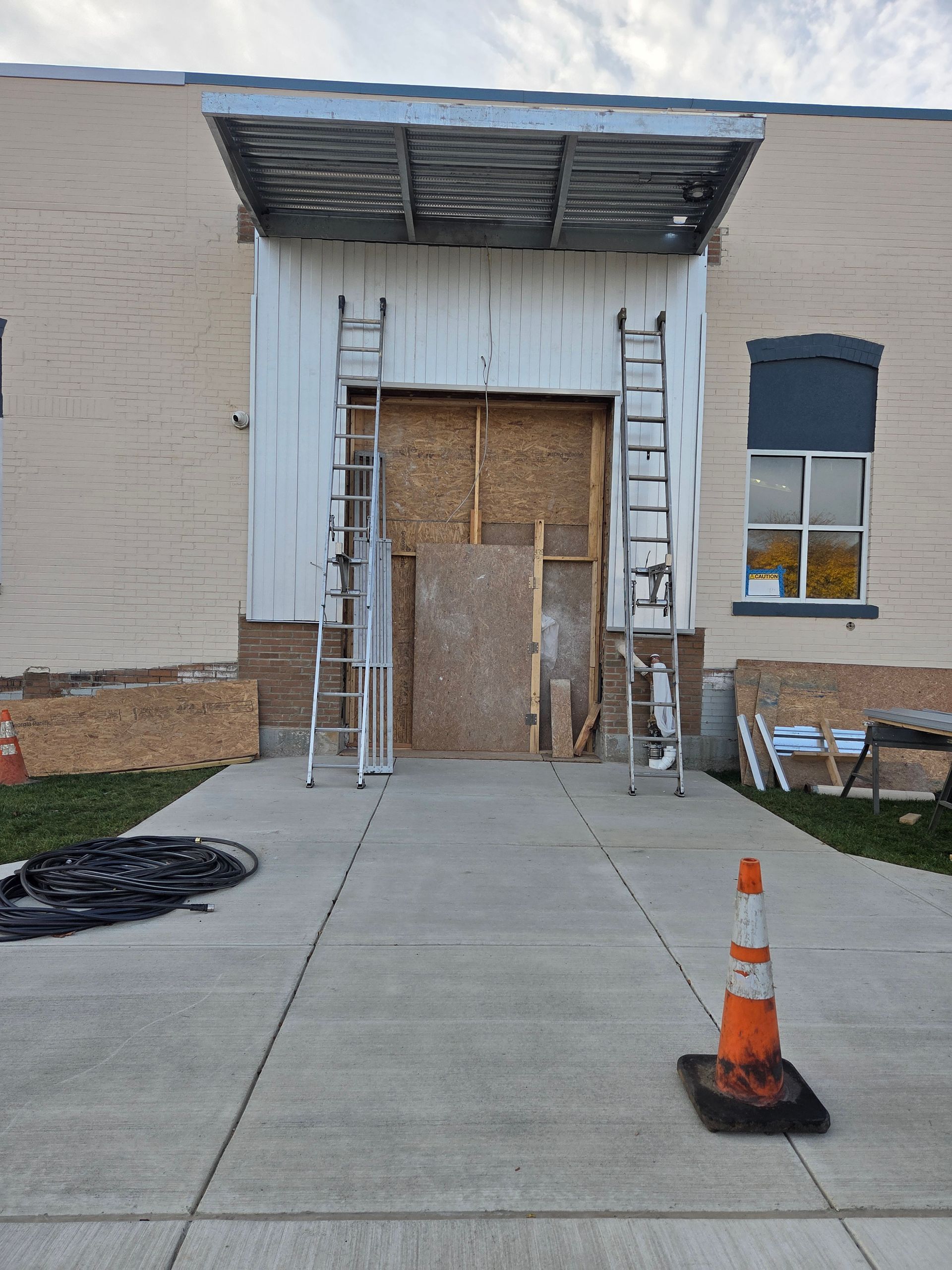 Construction site: door entry covered with plywood; ladders; safety cone; brick building; concrete walkway.