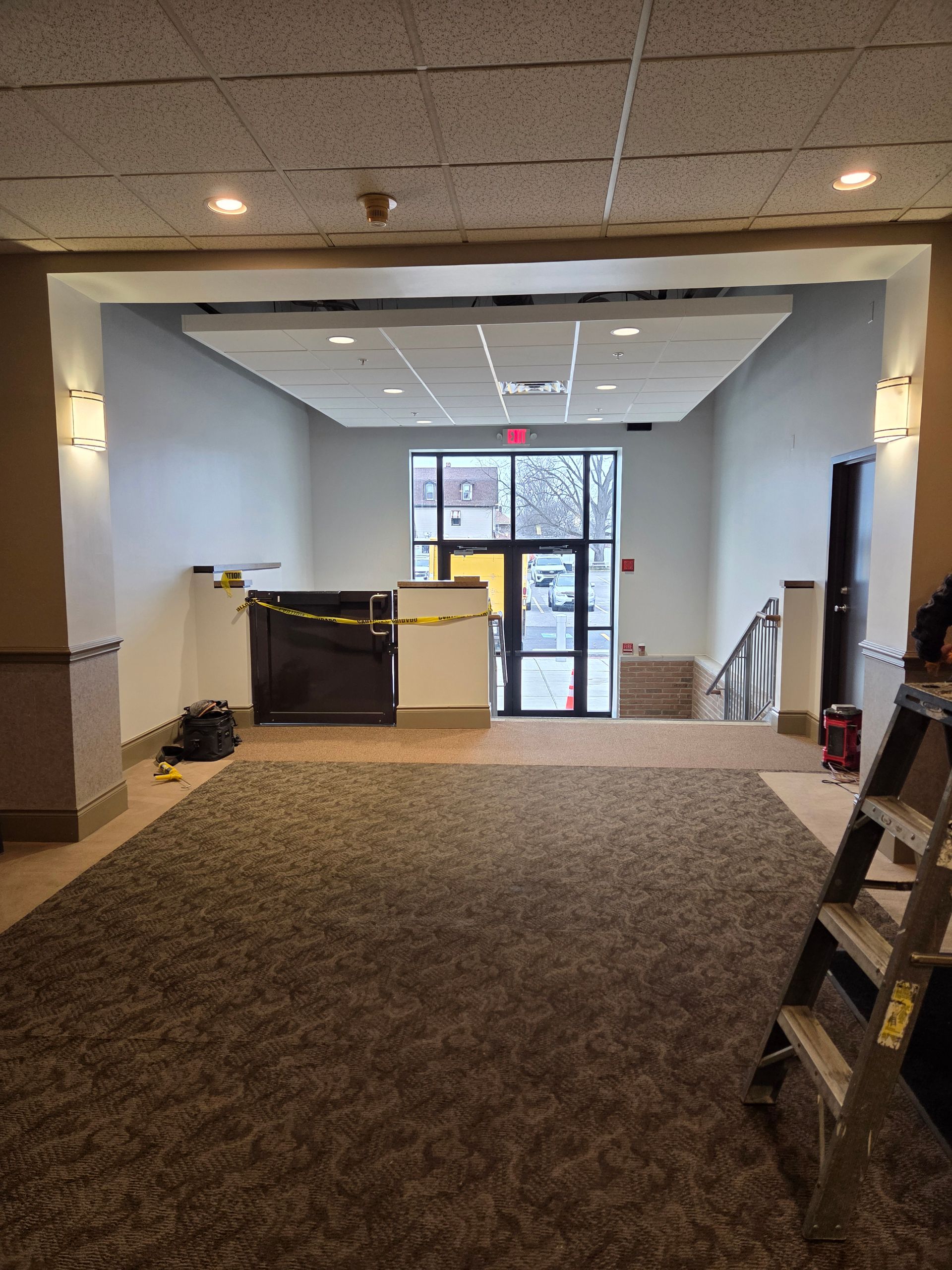 Hallway with brown carpet, open entrance with glass door, construction supplies visible.