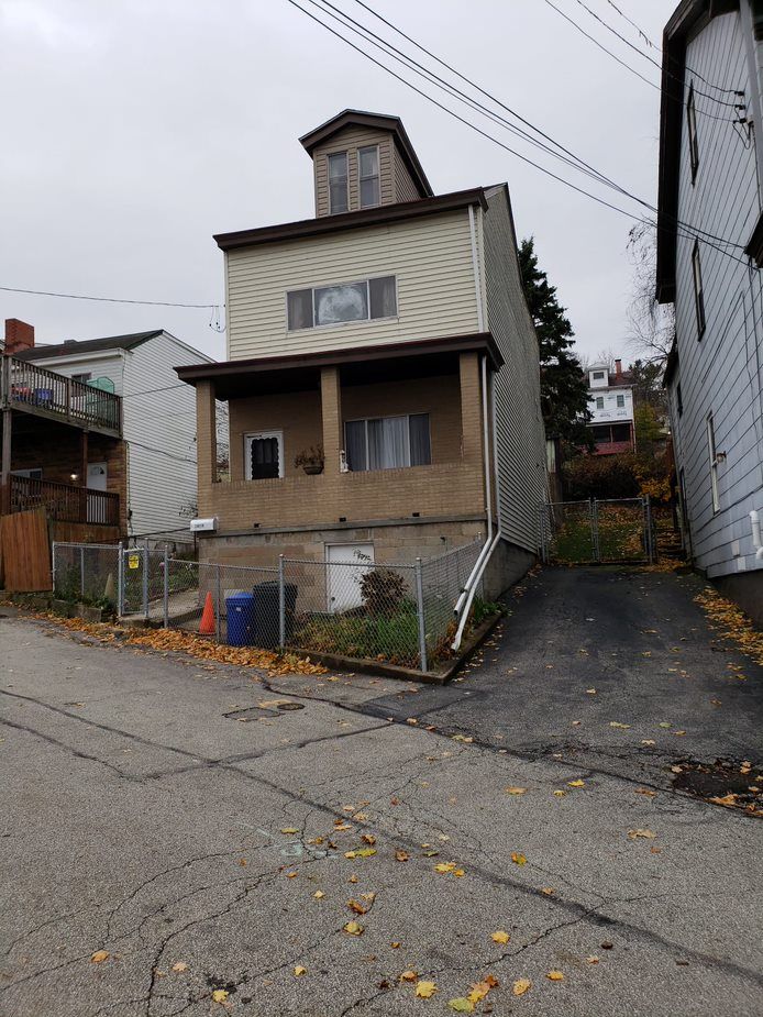 Three-story house on a sloped street; tan siding, brown roof, covered porch, trash cans in front.