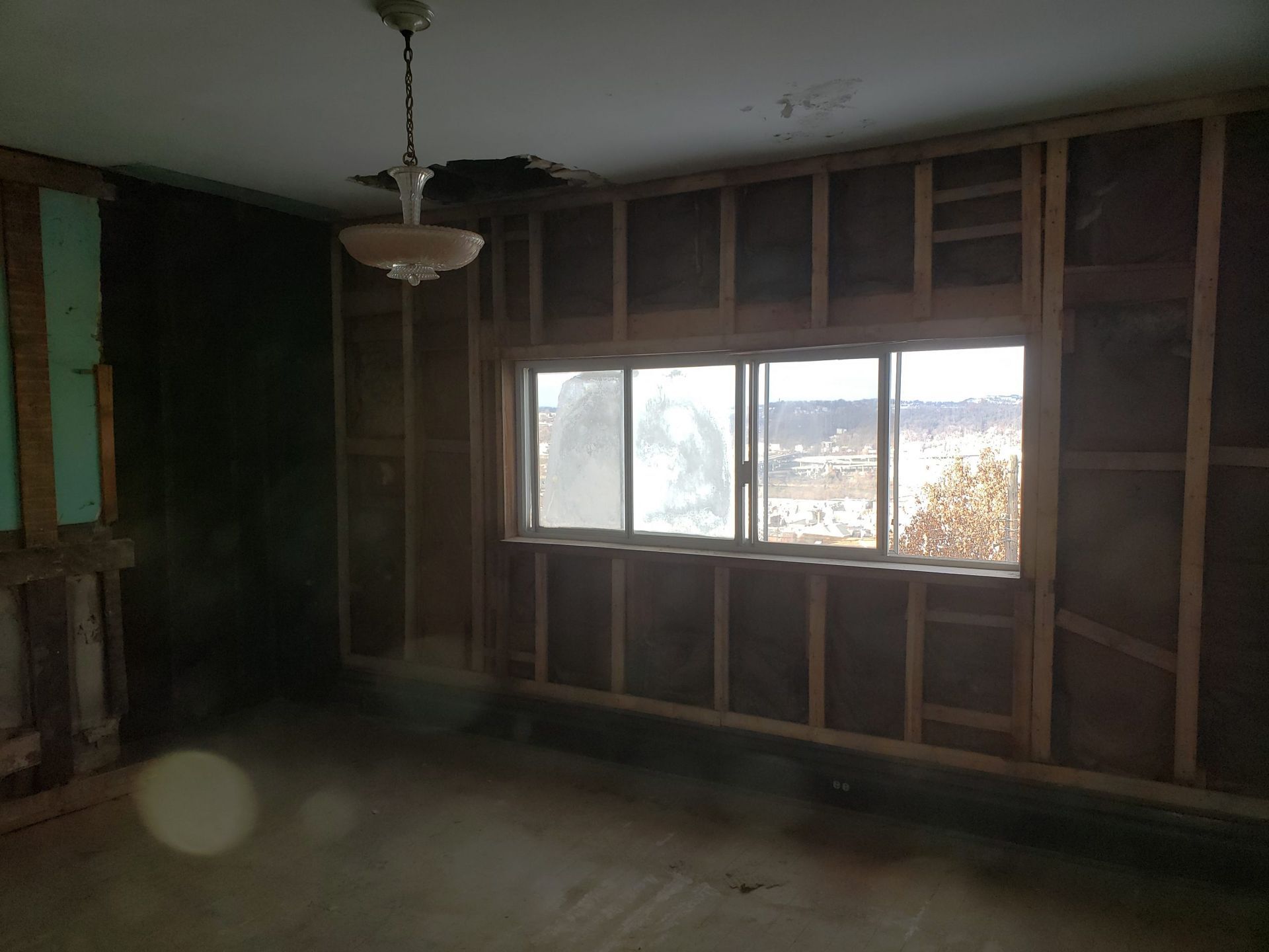 Interior room undergoing renovation, exposed studs, window, damaged ceiling, and hanging light fixture.