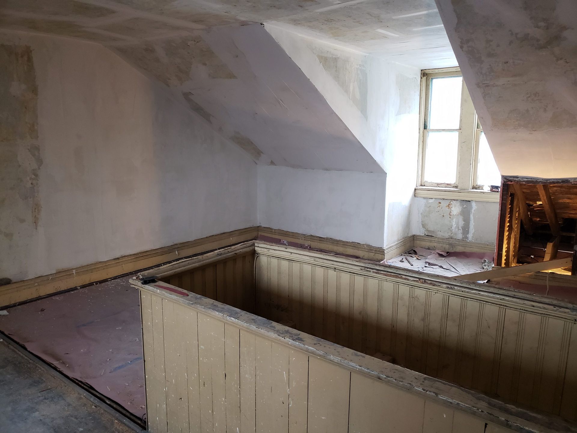 Interior of an attic room, with sloped walls, small window, and a wooden railing.