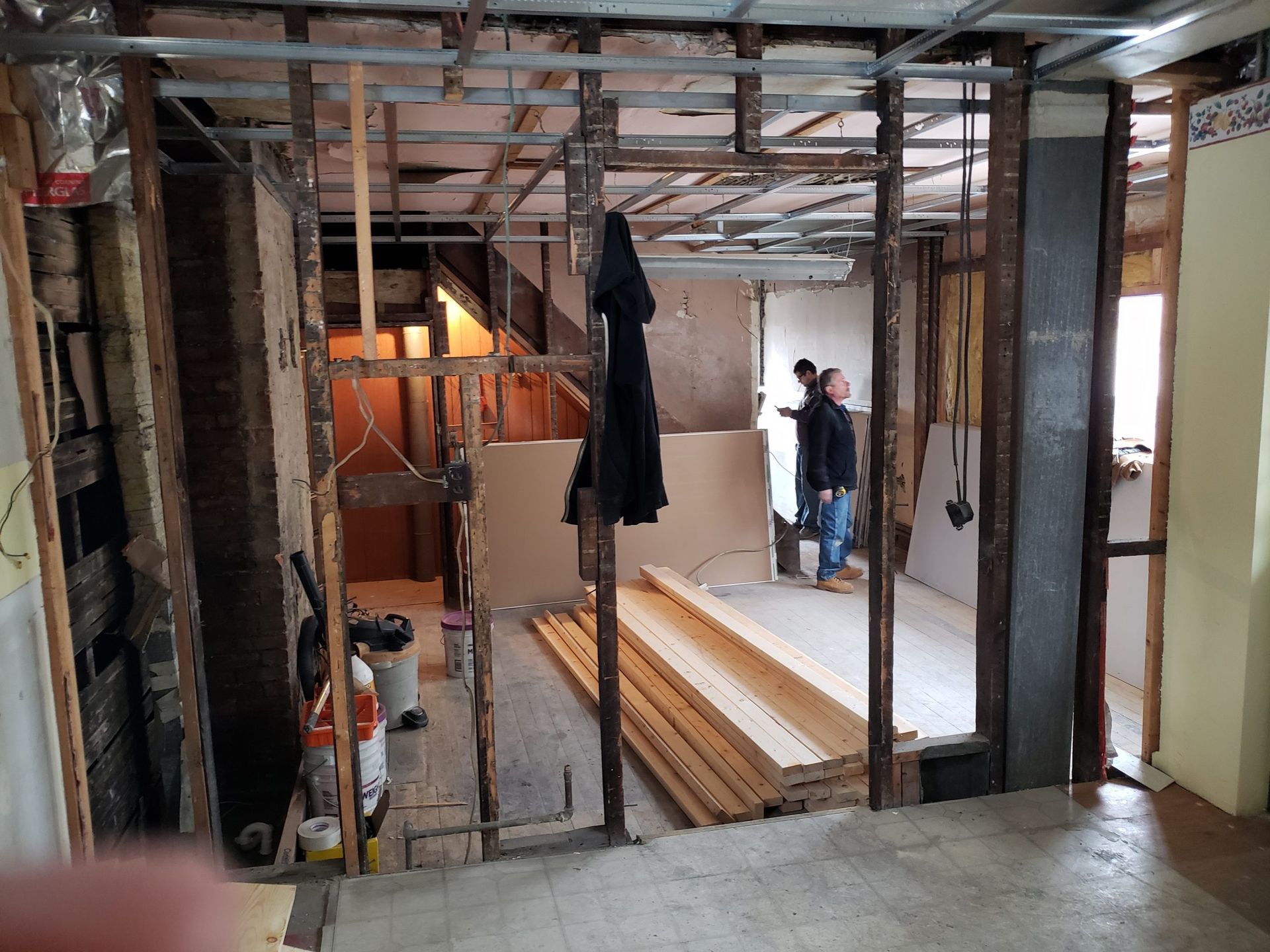 Interior renovation site: exposed framing, two people in the middle room, wood planks stacked.