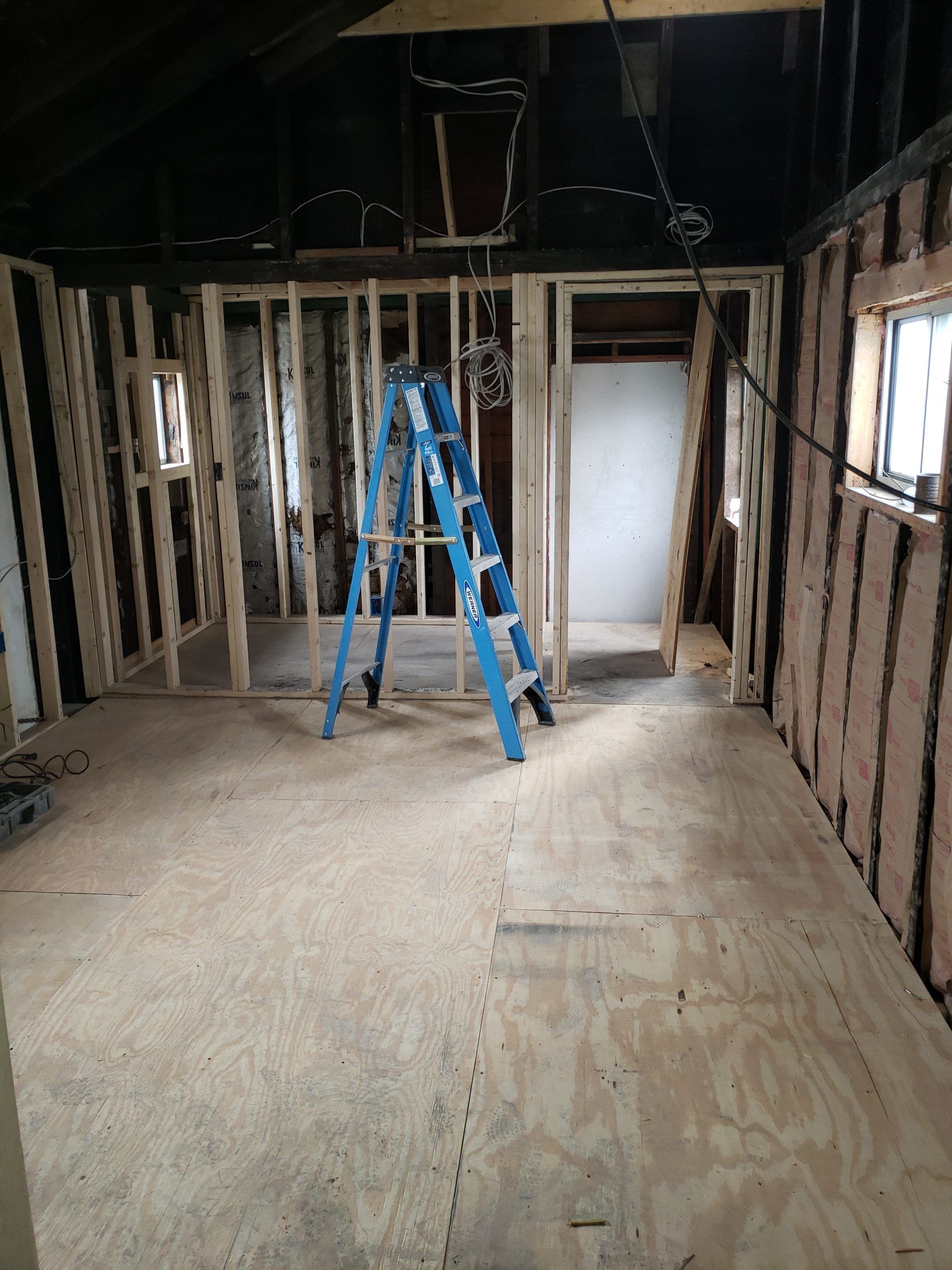 Interior room under construction with a blue ladder. Stud walls, plywood floor, and window frames are visible.