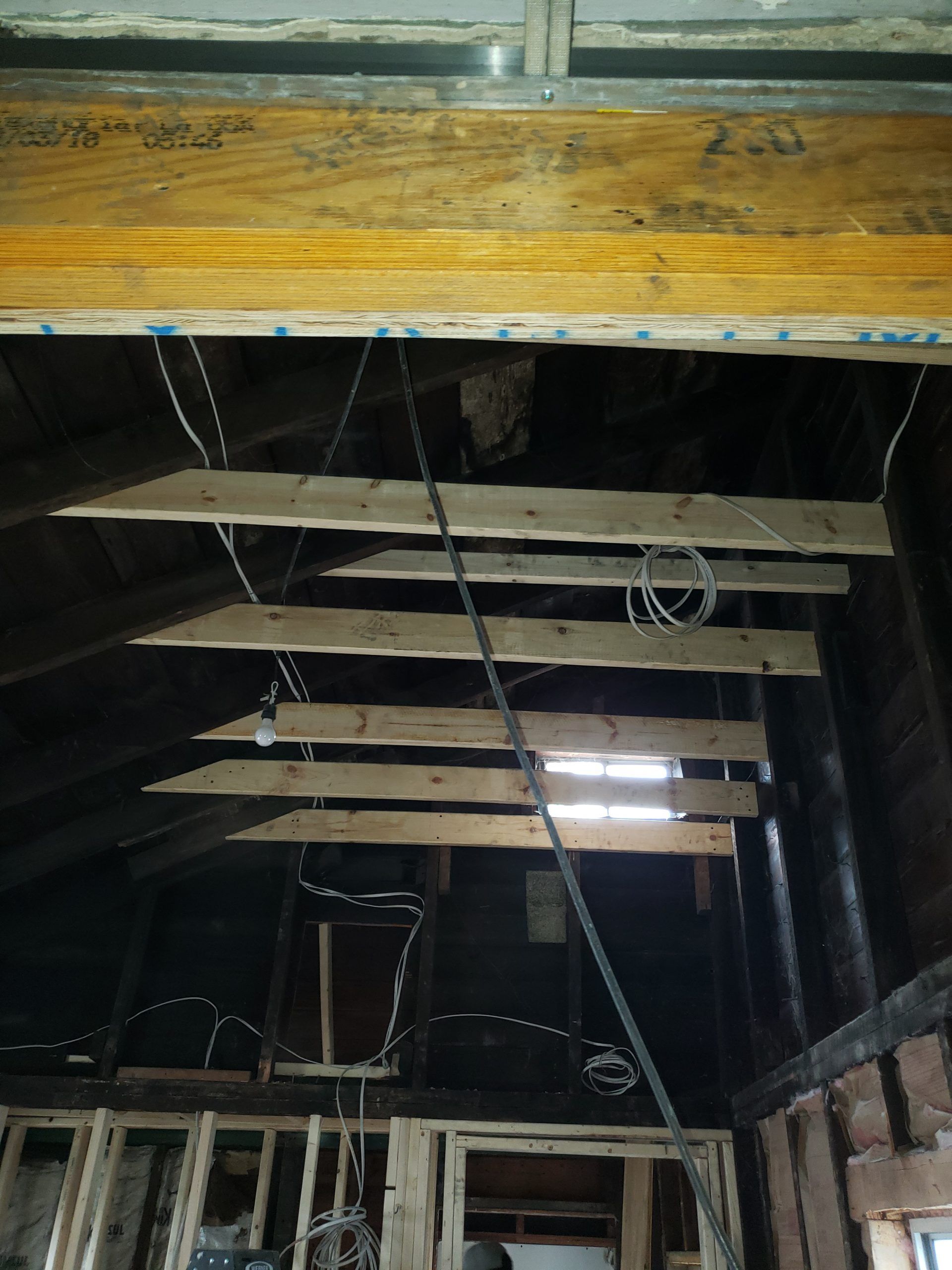 Interior view of a partially renovated attic ceiling with exposed wooden beams, wiring, and wooden planks.