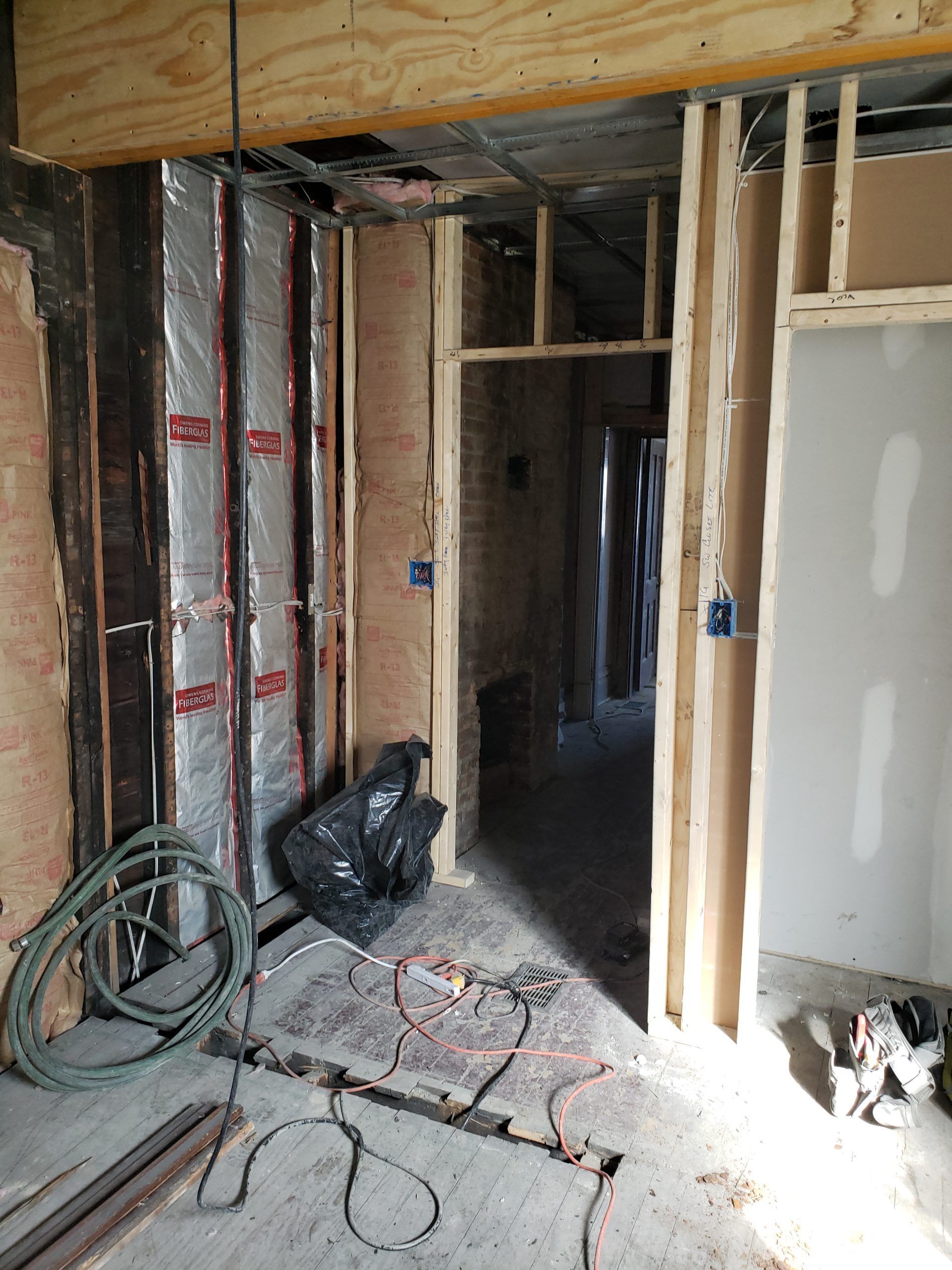 Interior renovation with exposed framing, insulation, and electrical wiring. A doorway leads to a hallway.