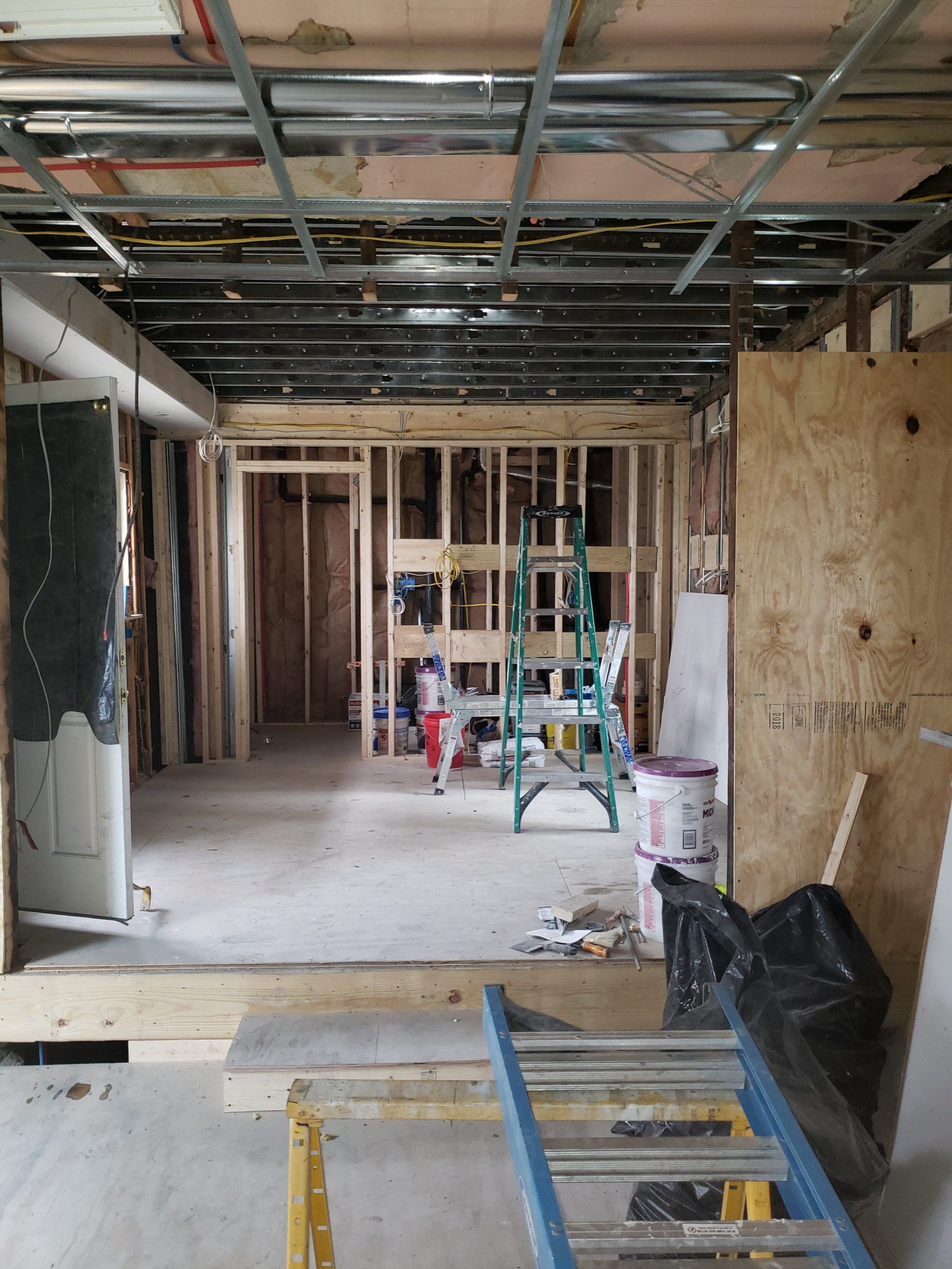 Interior of a building under construction, showing exposed wooden framing, ladder, and ceiling grid.