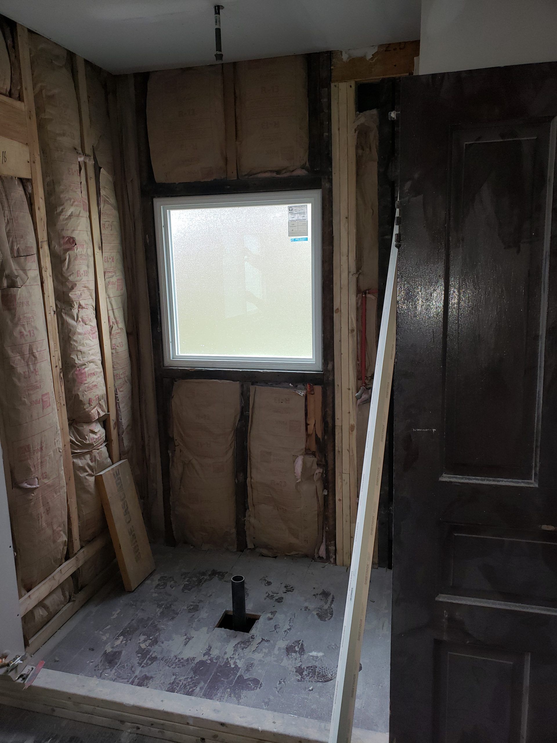 Bathroom renovation in progress, featuring a shower area with a window, exposed insulation, and a dark wooden door.