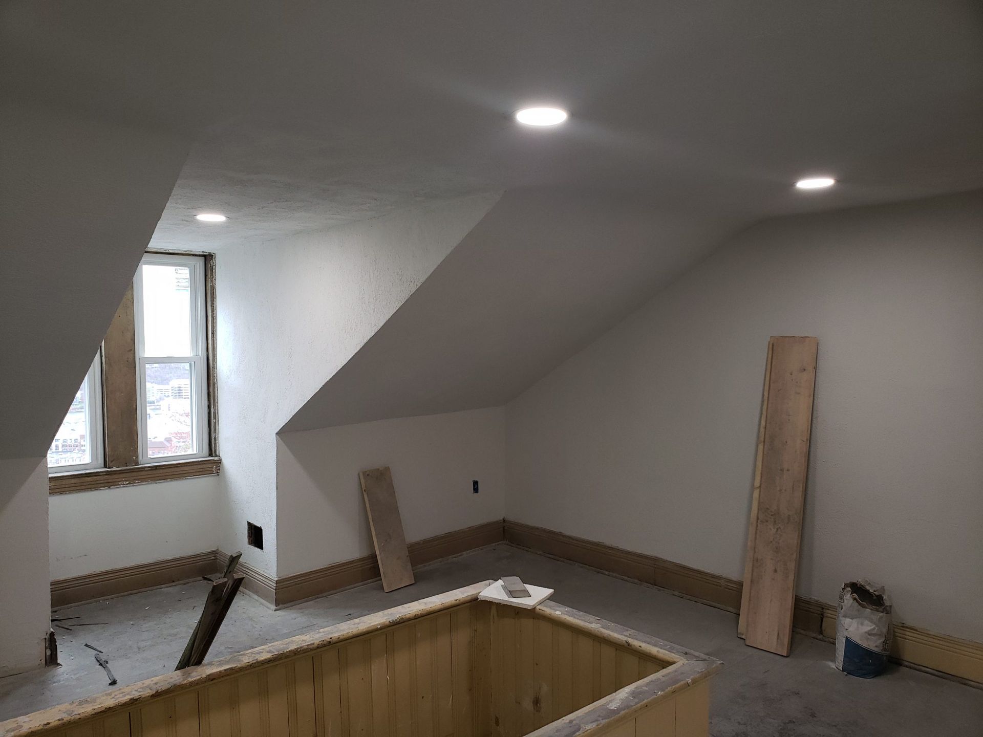Attic room under construction with sloped walls, window, recessed lights, and wooden trim.