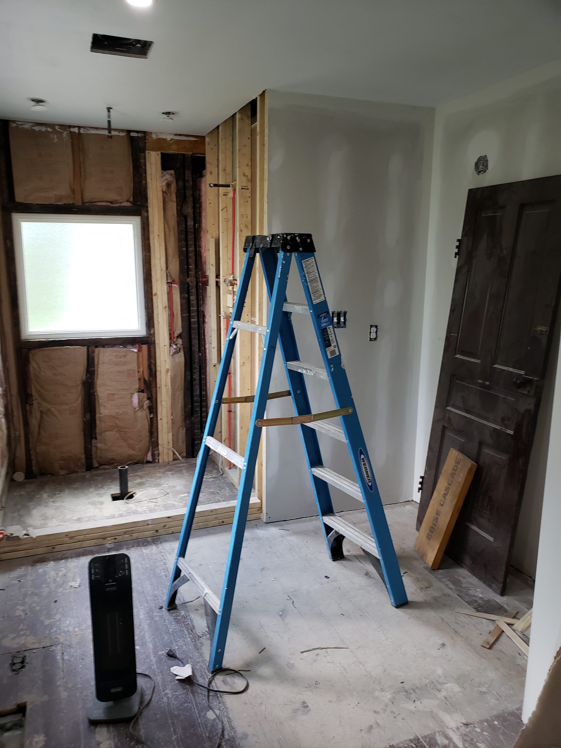 A blue ladder stands in a room under construction, beside drywall and a doorway.