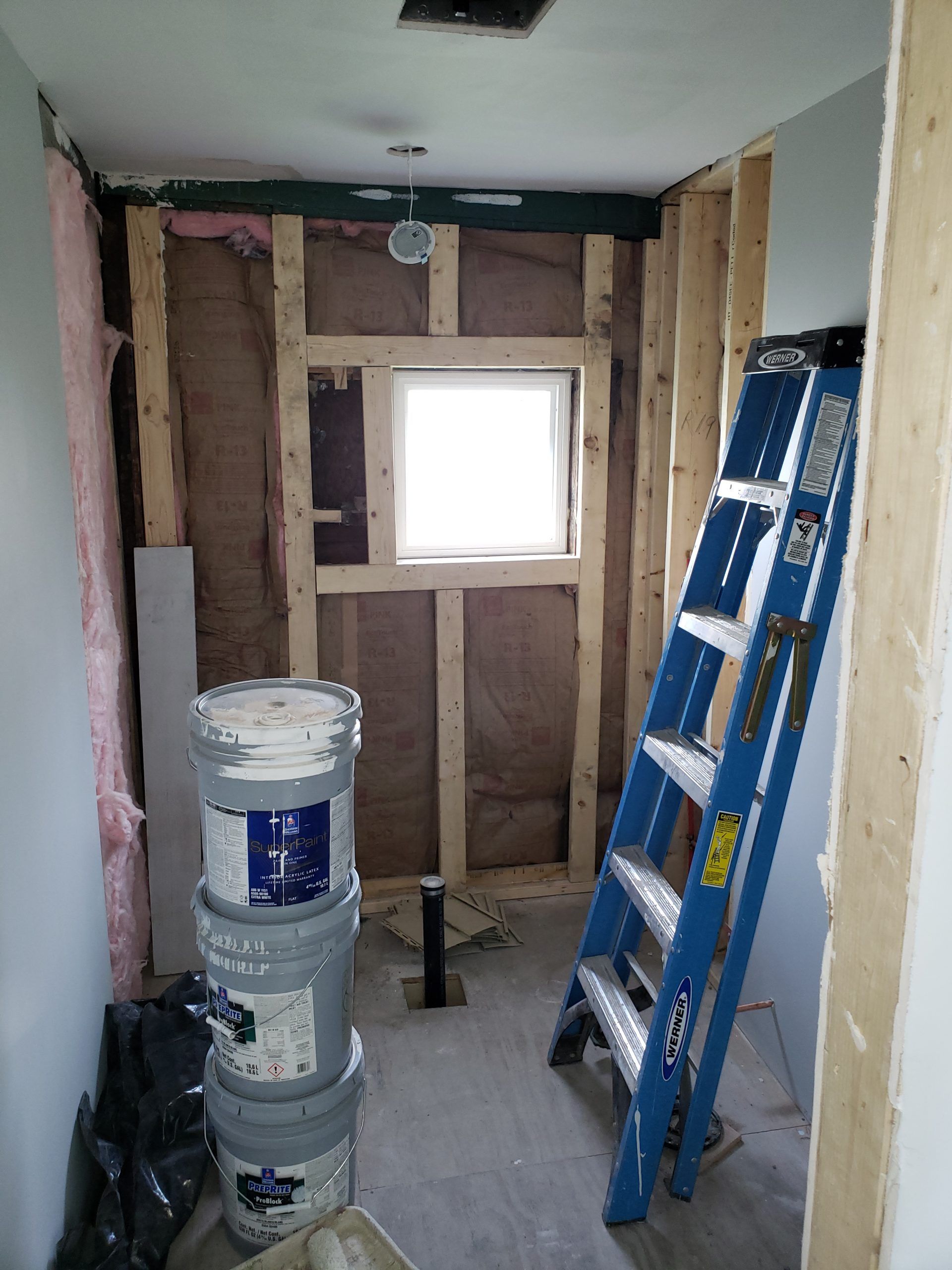 Bathroom under construction; framing, insulation, ladder, buckets, and window visible.
