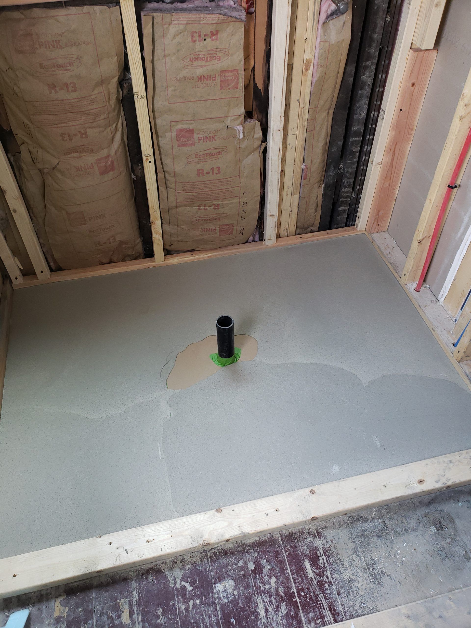 Shower stall under construction, with cement floor and drain pipe. Framing and insulation visible.