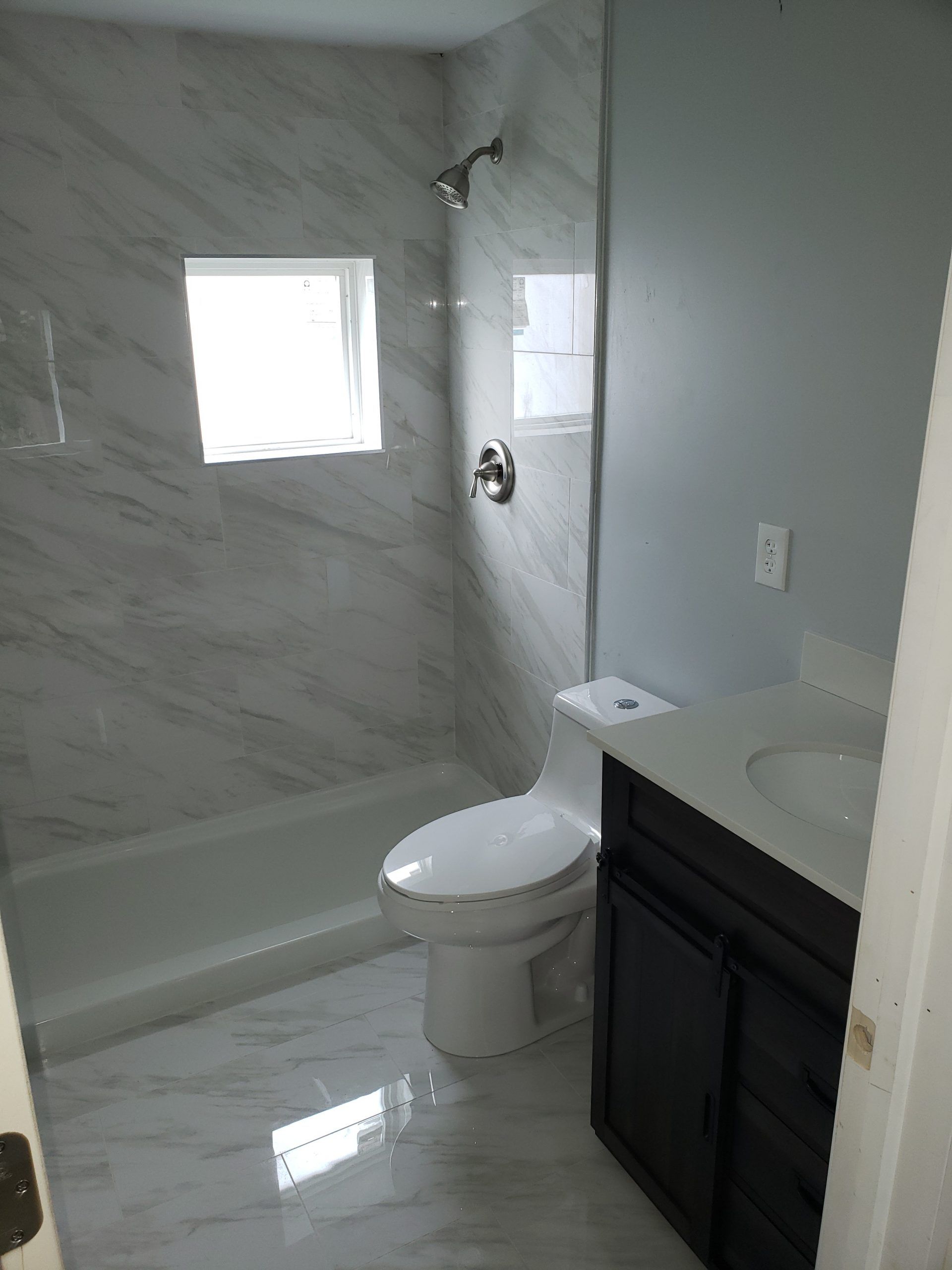 A remodeled bathroom with marble-look tile, a shower, a toilet, vanity, and a window.