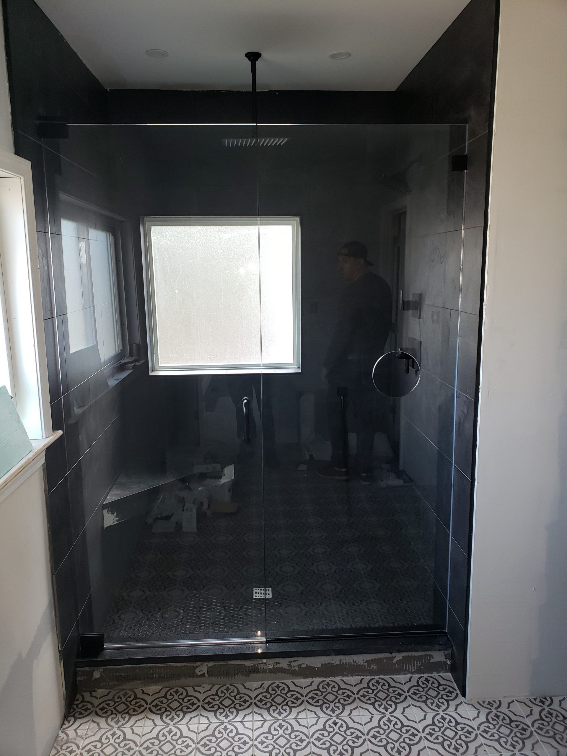 Shower with glass door, black tile walls, window, and patterned tile floor. Man inside.