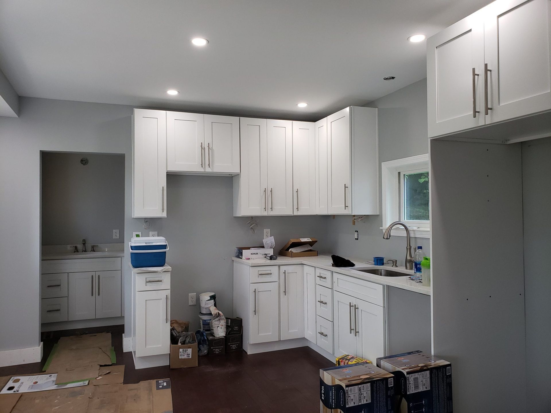 Newly installed white kitchen cabinets and sink in a room with gray walls and wood flooring.