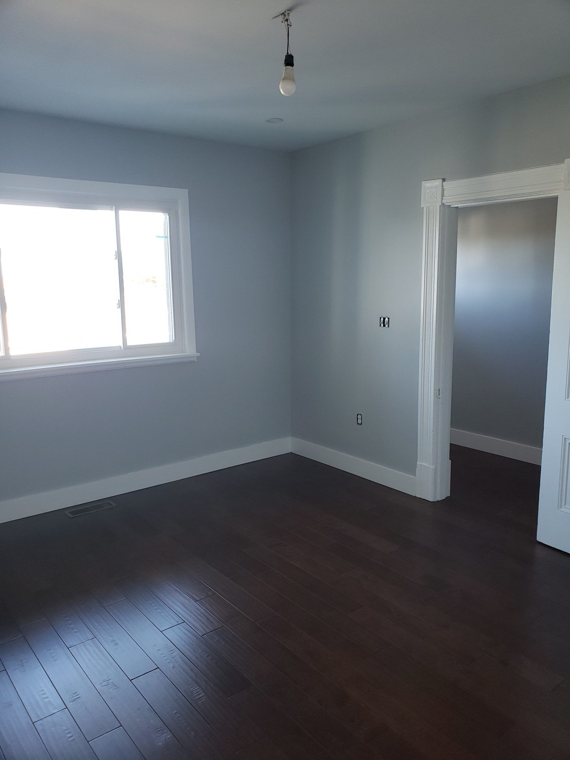 Empty room with light blue walls, dark wood floor, white trim, and a window.