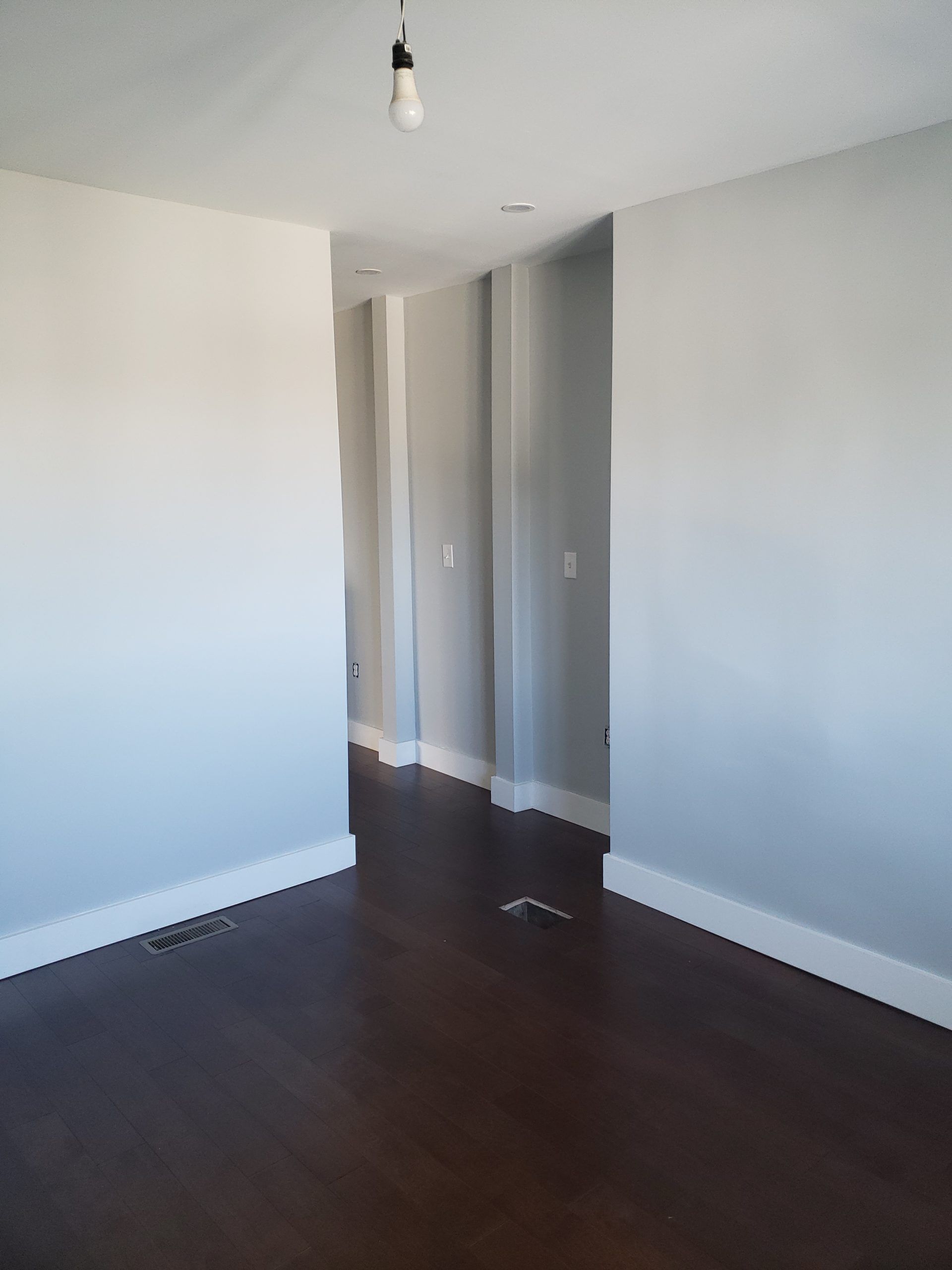 Empty room with light gray walls, dark wood floors, and a hallway leading to other rooms.