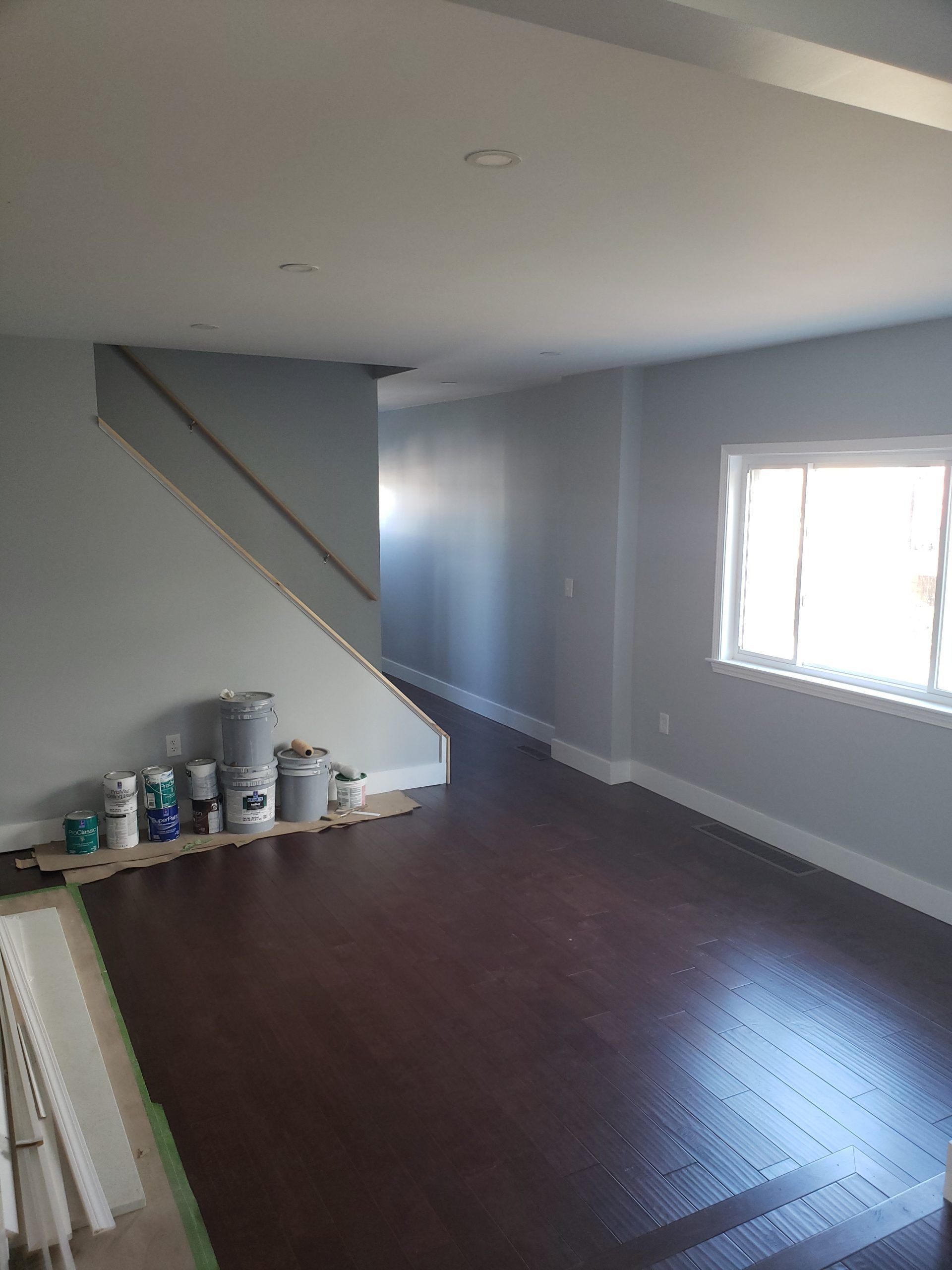 Room with hardwood floor, light blue walls, staircase, and window; paint cans sit on the floor.