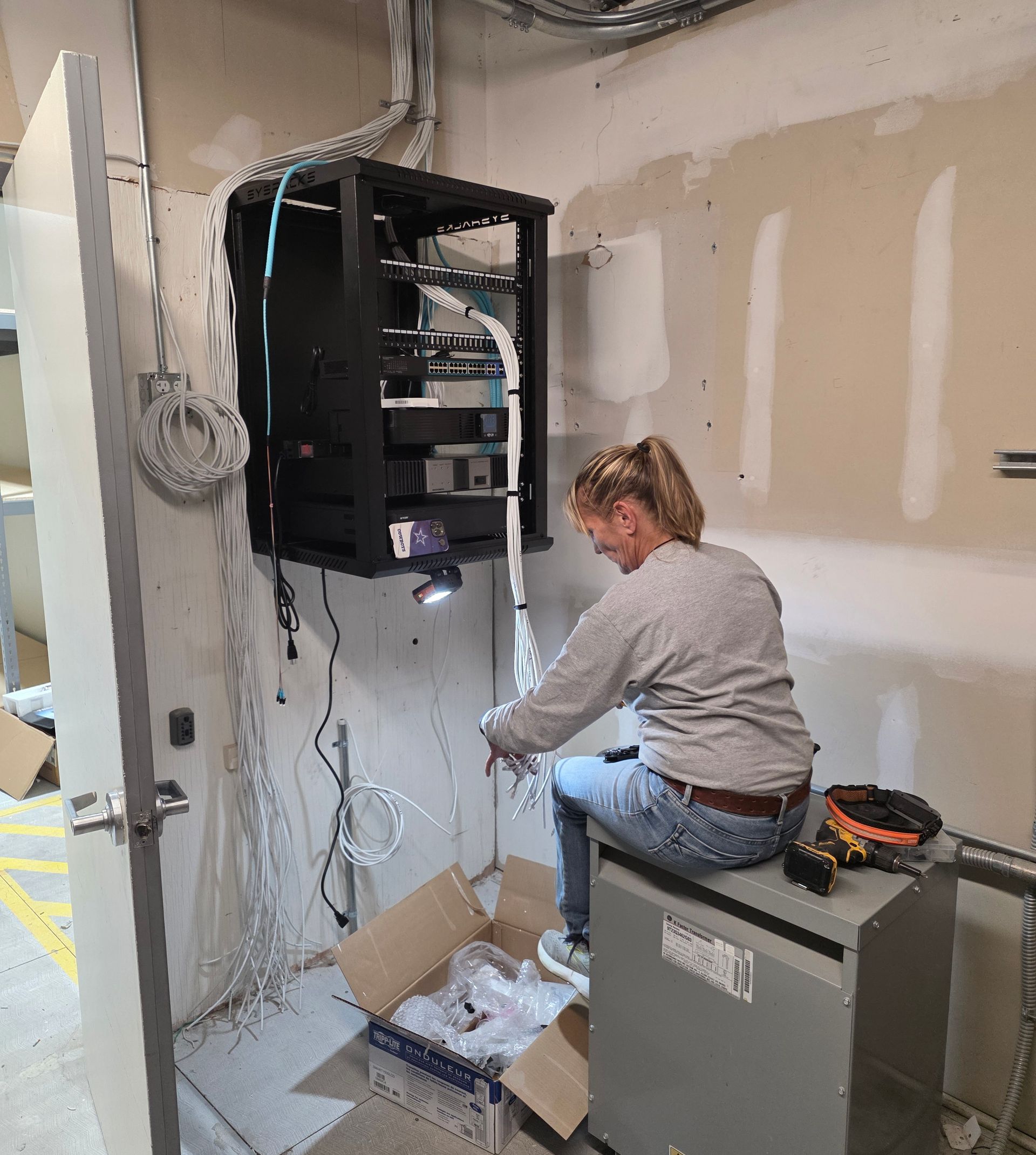 Technician kneeling beside an open wall-mounted network cabinet during an installation in a utility room