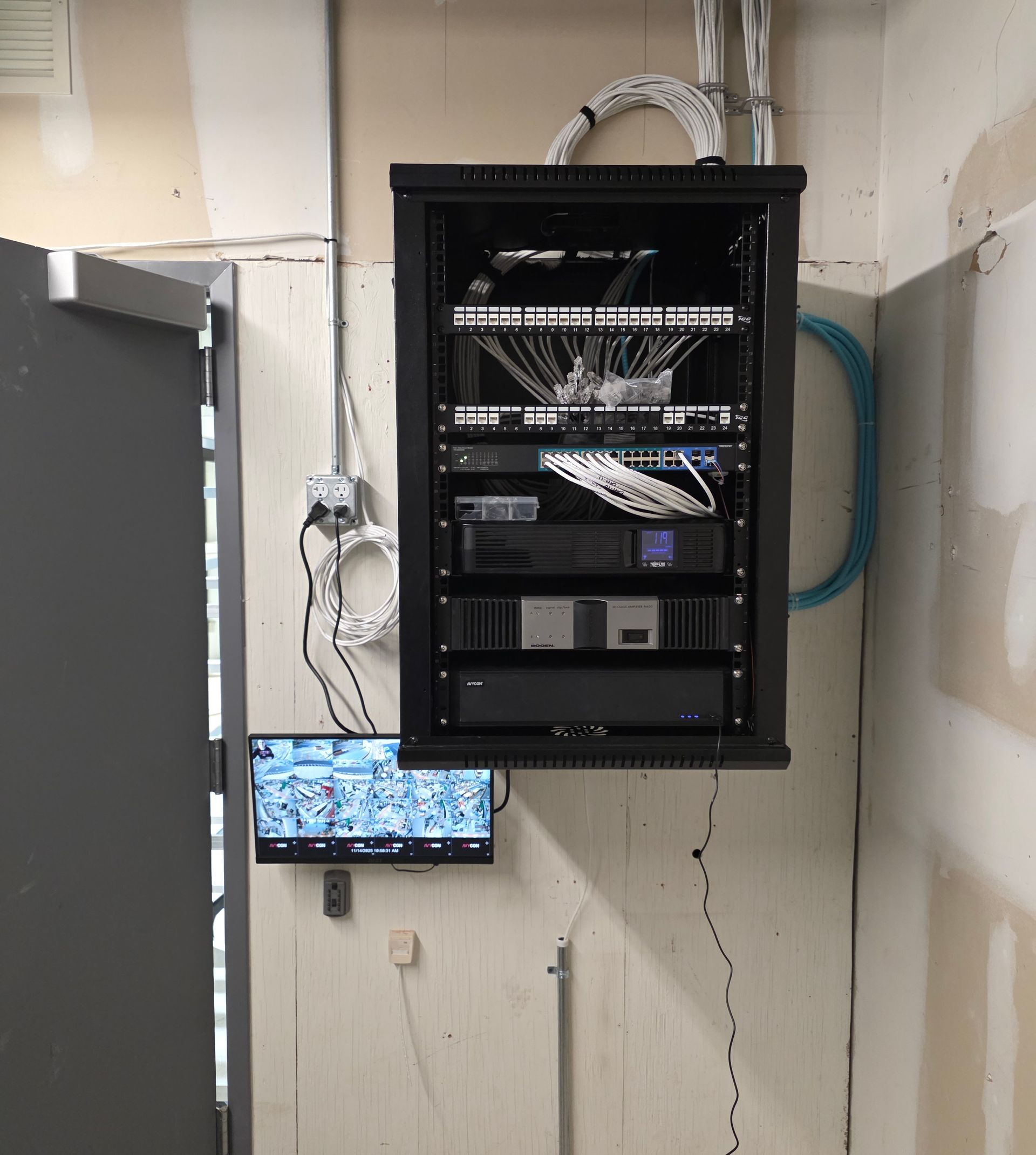 Wall-mounted network cabinet with cables and equipment above a small monitor in a utility room