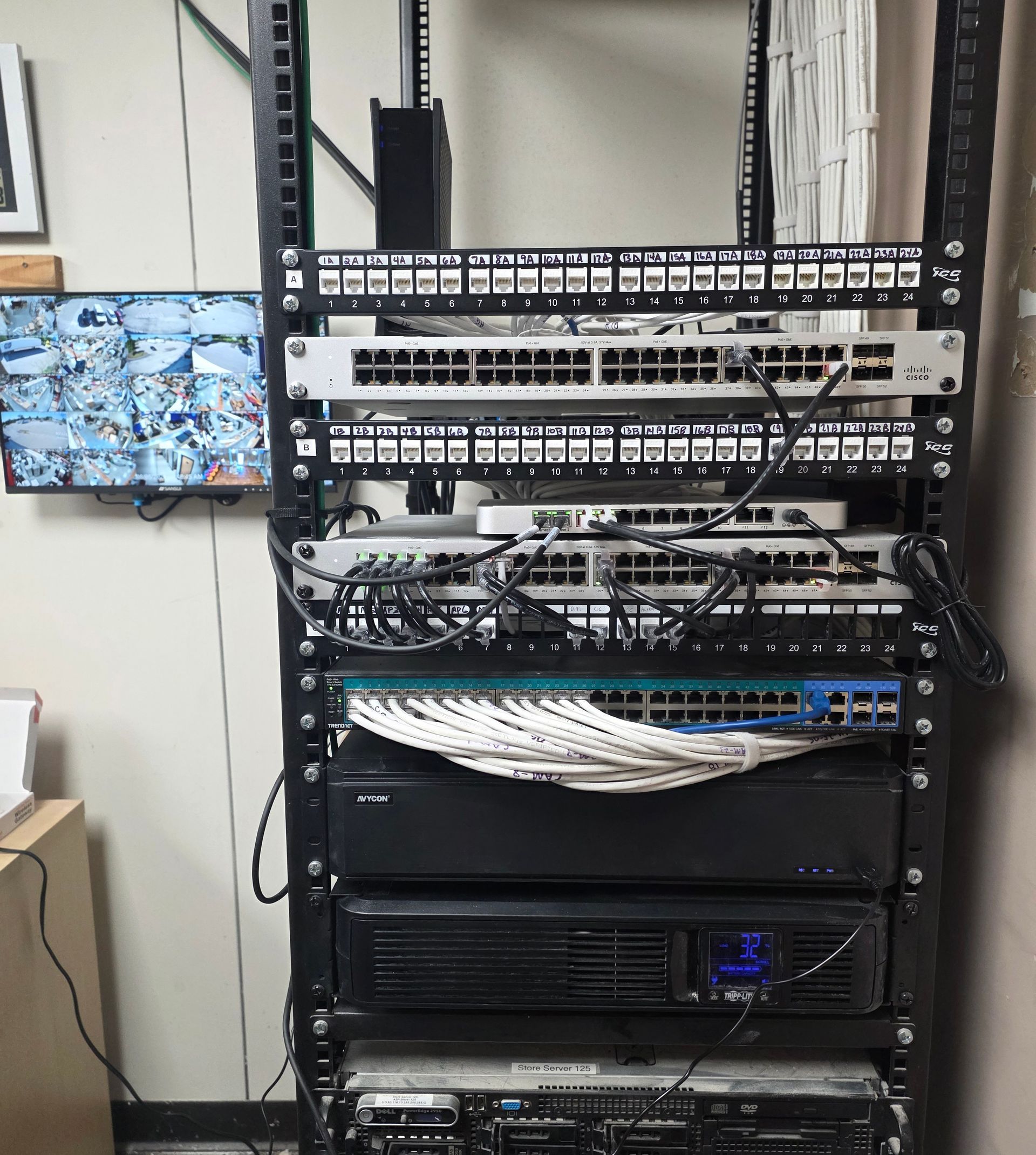 Network rack with stacked switches, routers, and cables beside a wall-mounted monitor.