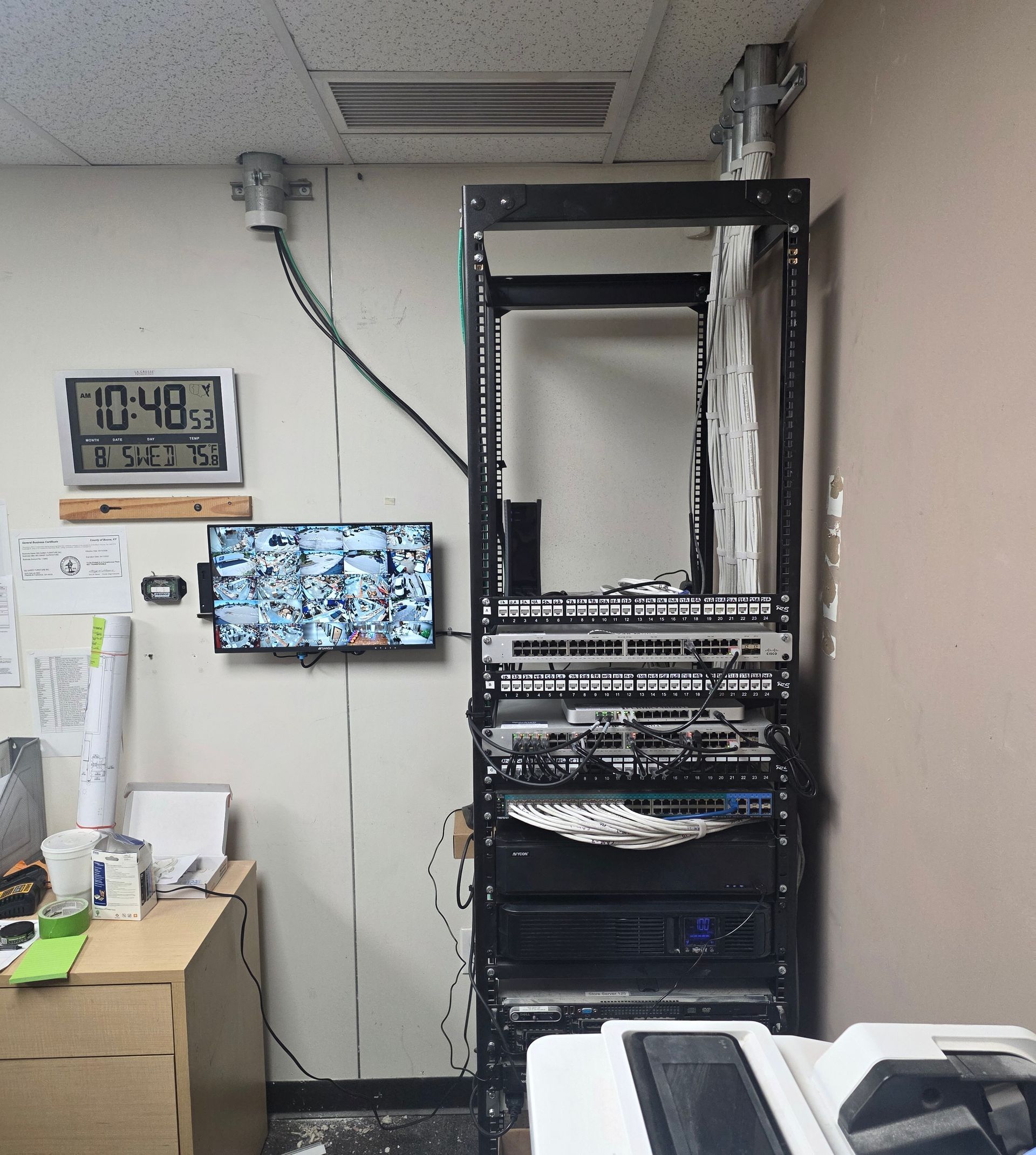 Server rack in an office corner with cables, wall clock, and desk items nearby