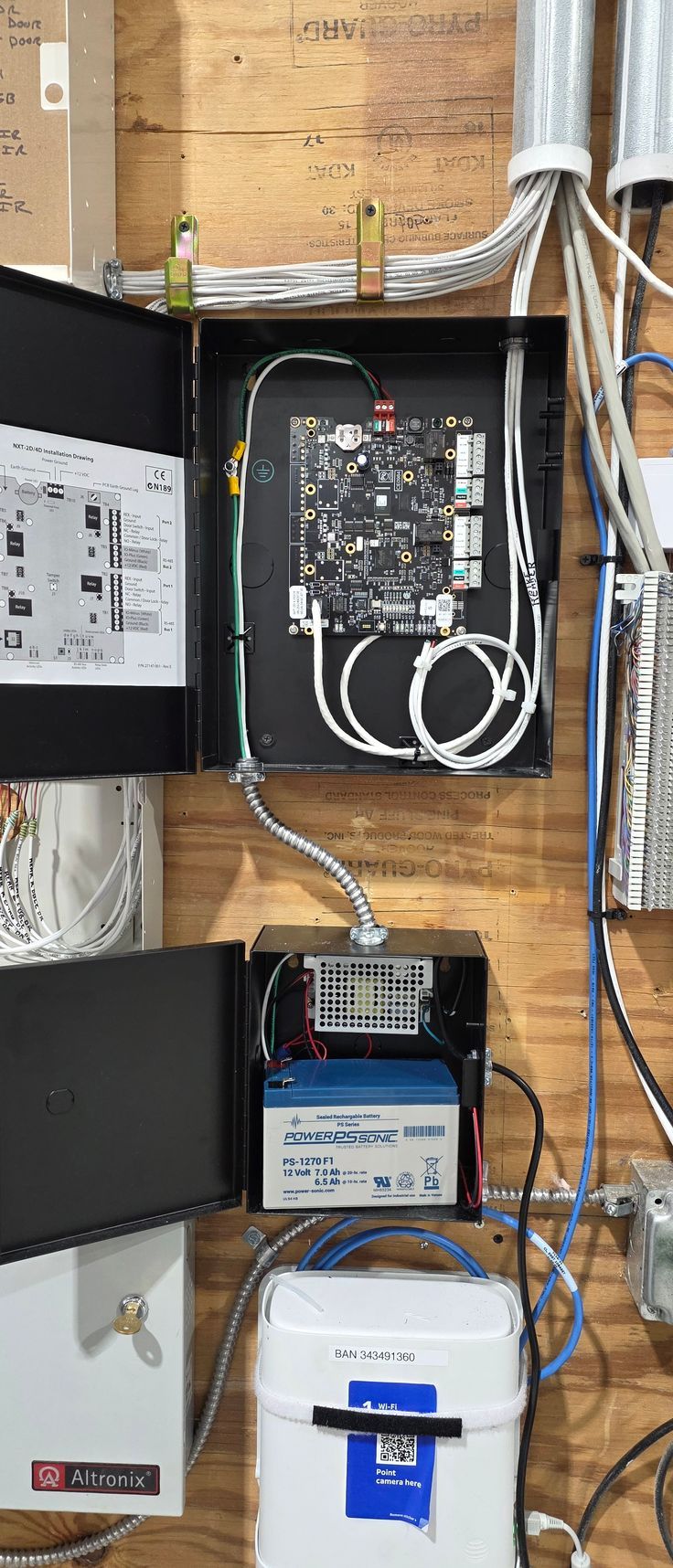 Open electrical panel with bundled cables and utility boxes mounted on a wood wall.