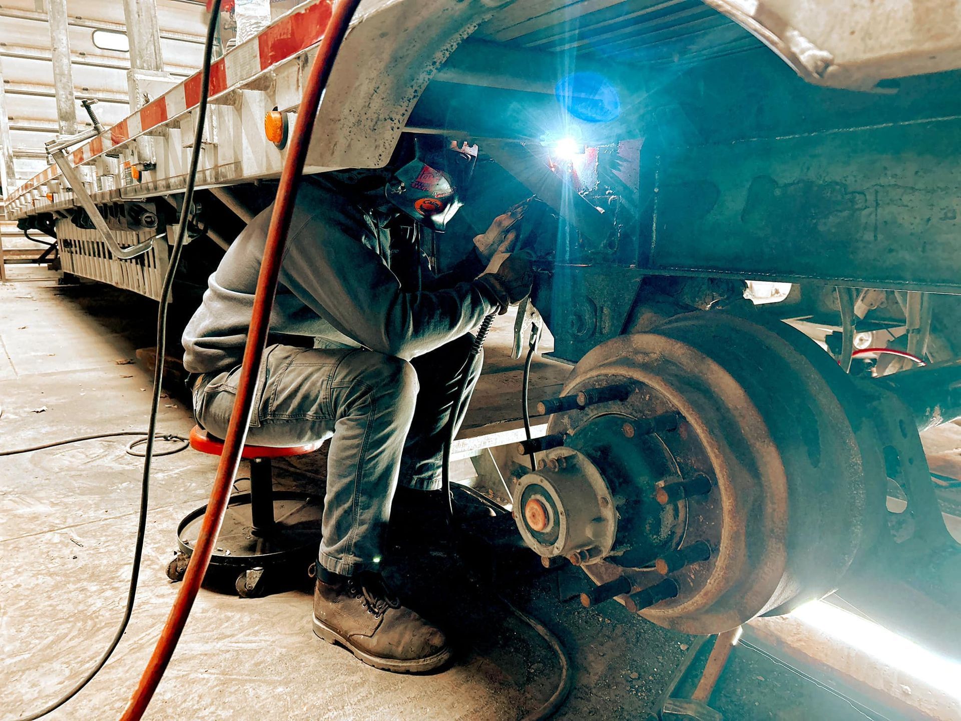 Welder wearing a mask working on the undercarriage of a trailer near a wheel.
