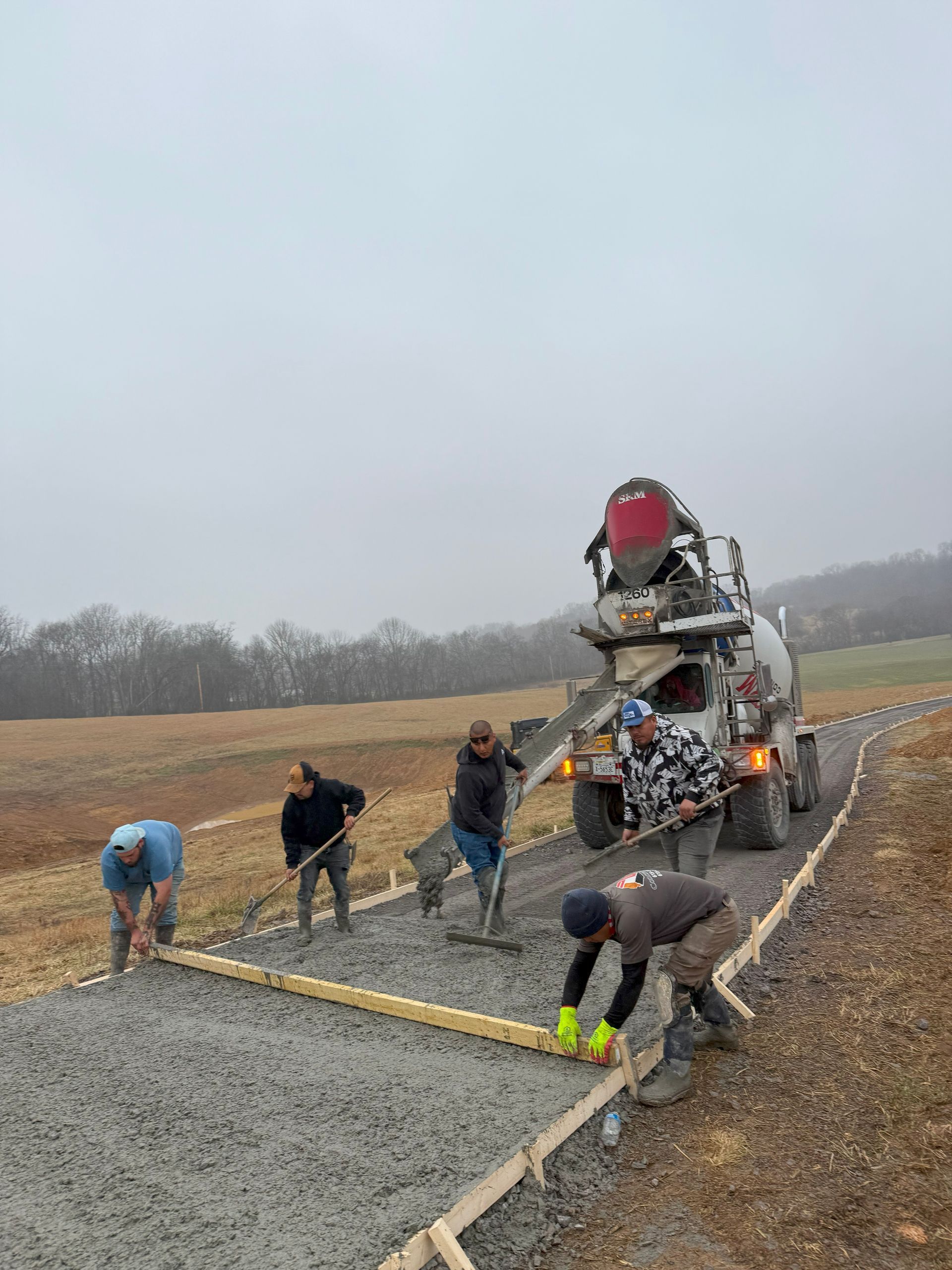 Workers pouring concrete from a mixer truck, leveling a path with tools in an open field on an overcast day.