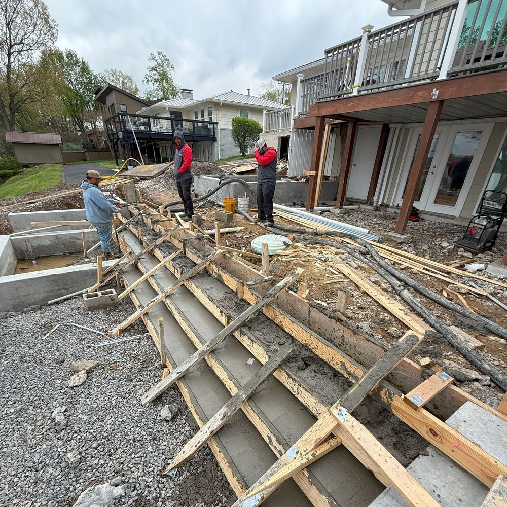 Construction workers building concrete steps near a house; wooden forms visible. Cloudy sky.