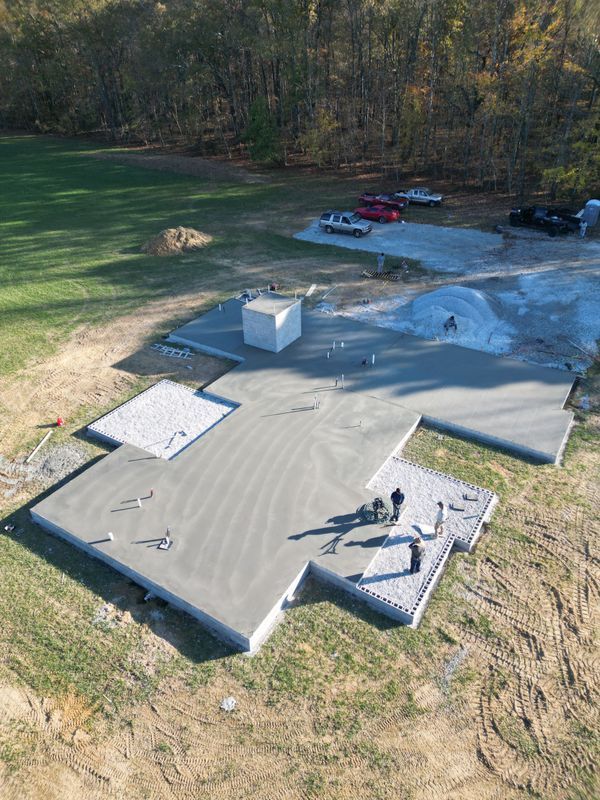 Newly poured concrete foundation with workers; outdoors in a grassy area, vehicles in the background.