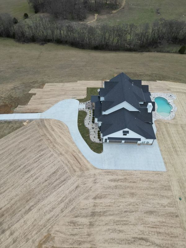 Aerial view of a large house with a black roof, long driveway, and a pool in a grassy field.