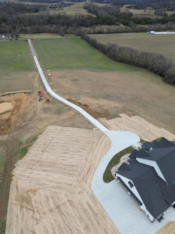Long concrete driveway winding through fields to a house; construction equipment visible.