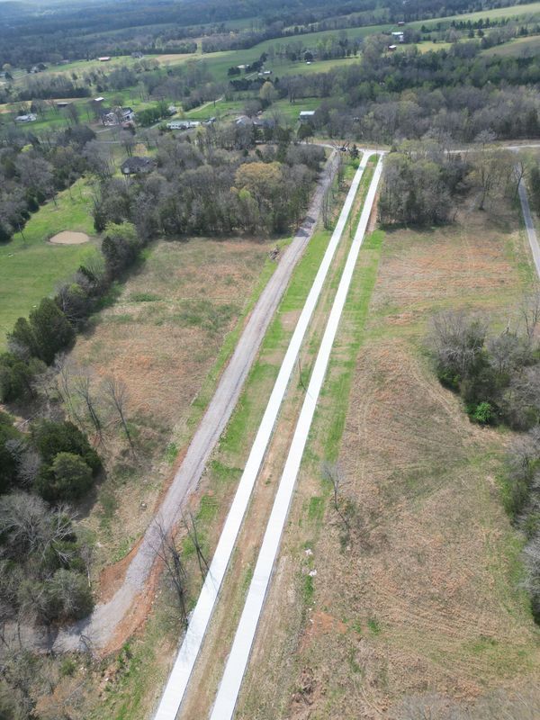 Aerial view of long, parallel, light-colored concrete paths through a rural, grassy area. Trees and fields surround.