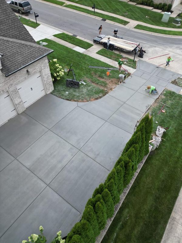 Newly poured concrete driveway with workers. Green grass, trees, and a house with a gray roof are visible.