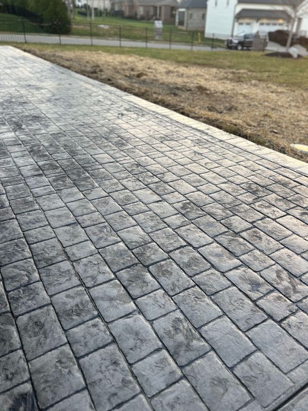 Concrete driveway with a brick pattern. Grey surface, grass and houses in the background.
