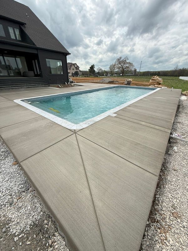 A rectangular swimming pool with concrete decking, next to a black house under an overcast sky.