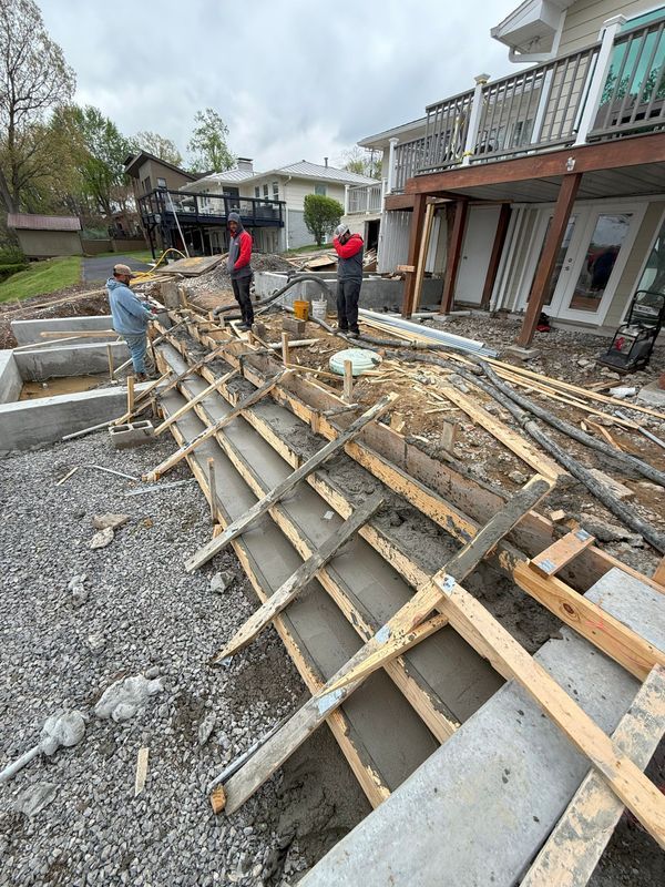 Construction workers pouring concrete into wooden forms on a deck, next to a house.